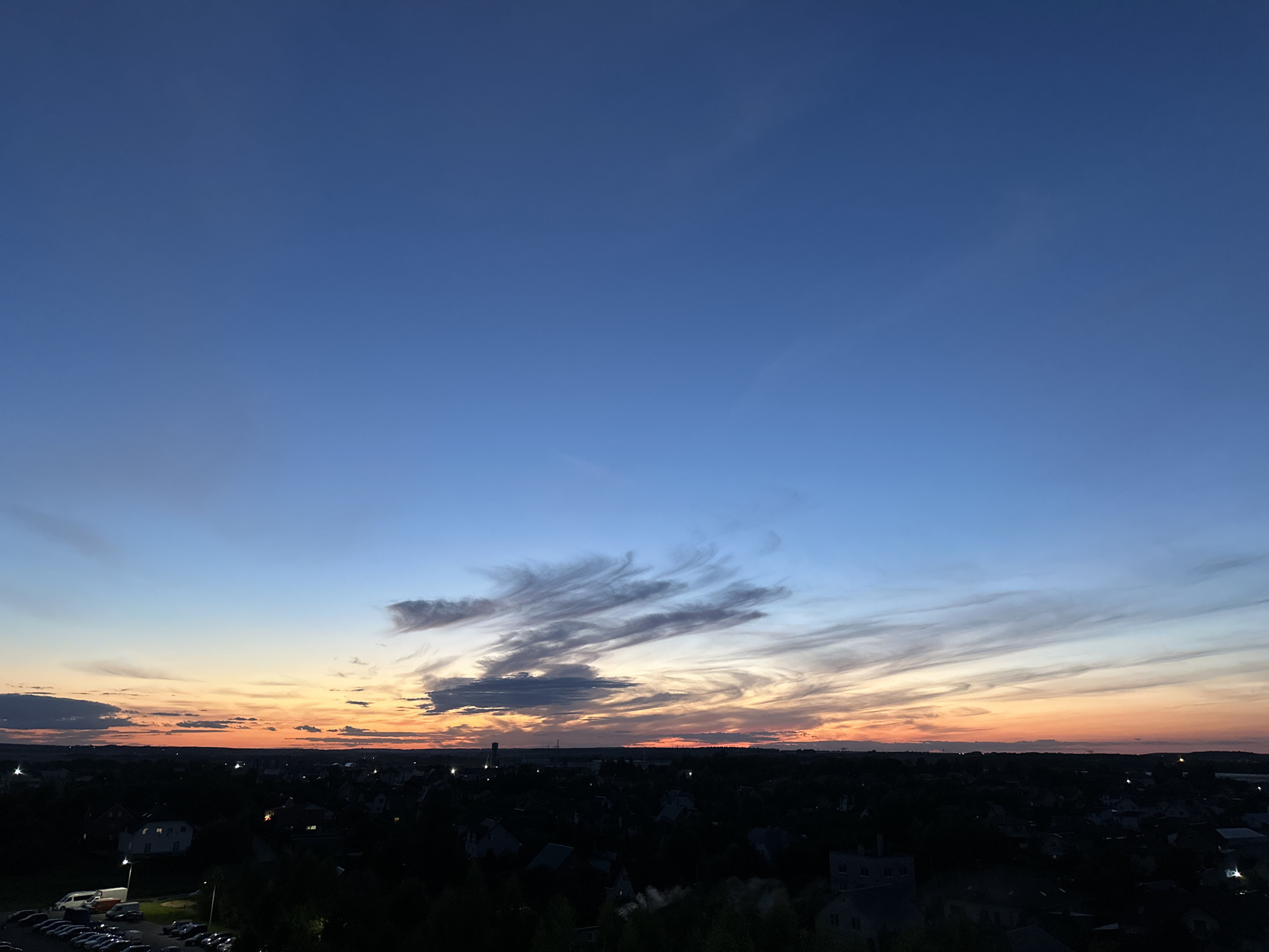 An aerial view captures the twilight sky over a city in Шчомысліцкі сельскі Савет, Belarus. The sky transitions from a deep blue at the top to hues of orange and yellow near the horizon, where wispy clouds are illuminated by the setting sun. Below, the silhouette of a town is visible, with dark trees and rooftops dotted with lights. In the lower left corner, a parking lot with several vehicles is discernible. The scene is devoid of any visible people or active events.