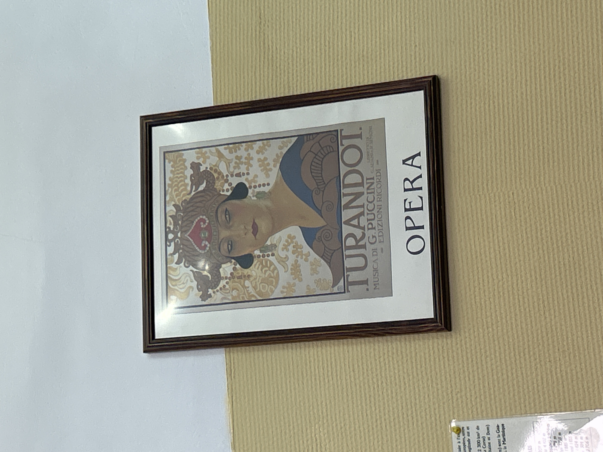 A framed poster of "Turandot" opera by G. Puccini hangs on a wall. The wall is divided into two sections, the left is white and the right is beige with a ribbed texture. A piece of paper with text is visible at the bottom right.