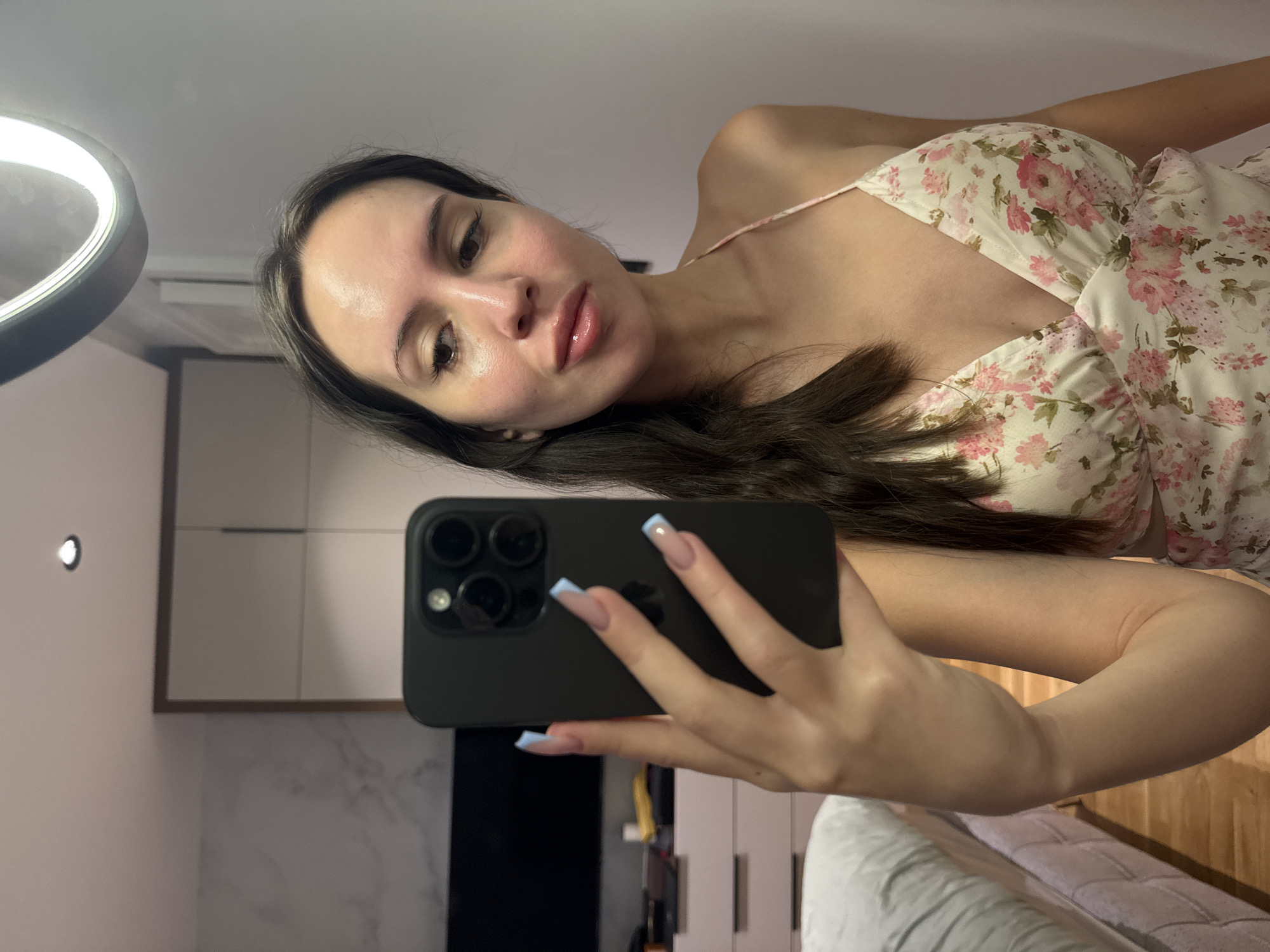 A woman is taking a selfie in a bedroom. She is wearing a floral-print dress and has long, dark hair. She is holding a black smartphone in her left hand. The room appears to be furnished with a bed, a dresser, and a closet. There is also a ring light visible in the upper right corner of the image.