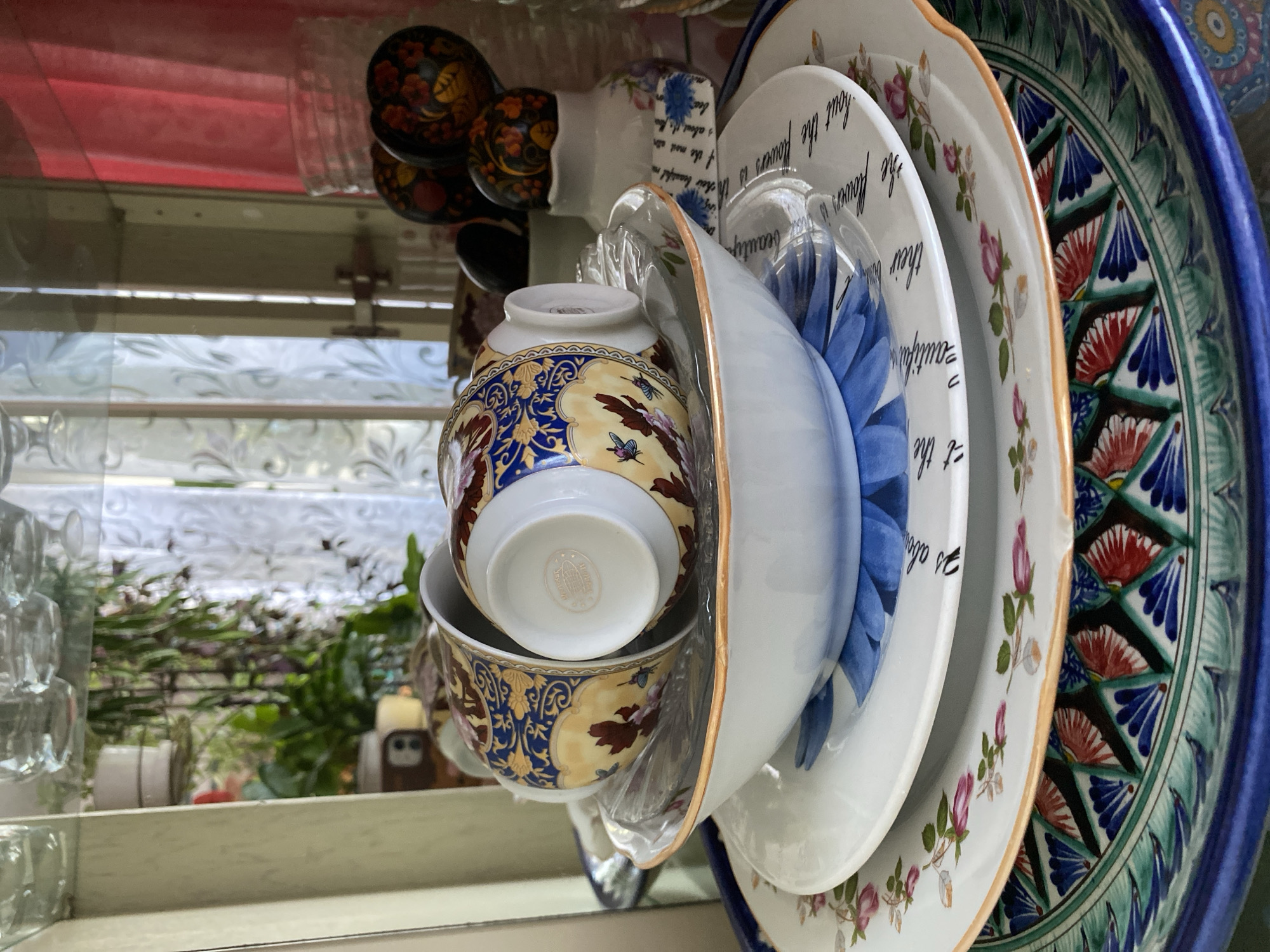 The image shows a stack of decorative plates and teacups. The plates feature various floral patterns and designs, including blue and red fan-like shapes on a larger, outermost plate, and smaller pink roses on another. One plate has text on it. Two teacups, decorated with yellow and blue designs and butterflies, are stacked one inside the other. The setting appears to be a shelf or display area, possibly inside a home or shop, with a window visible in the background. Outside the window, greenery and other objects are visible. The image does not include information to verify the location in Votkinsk, Russia.