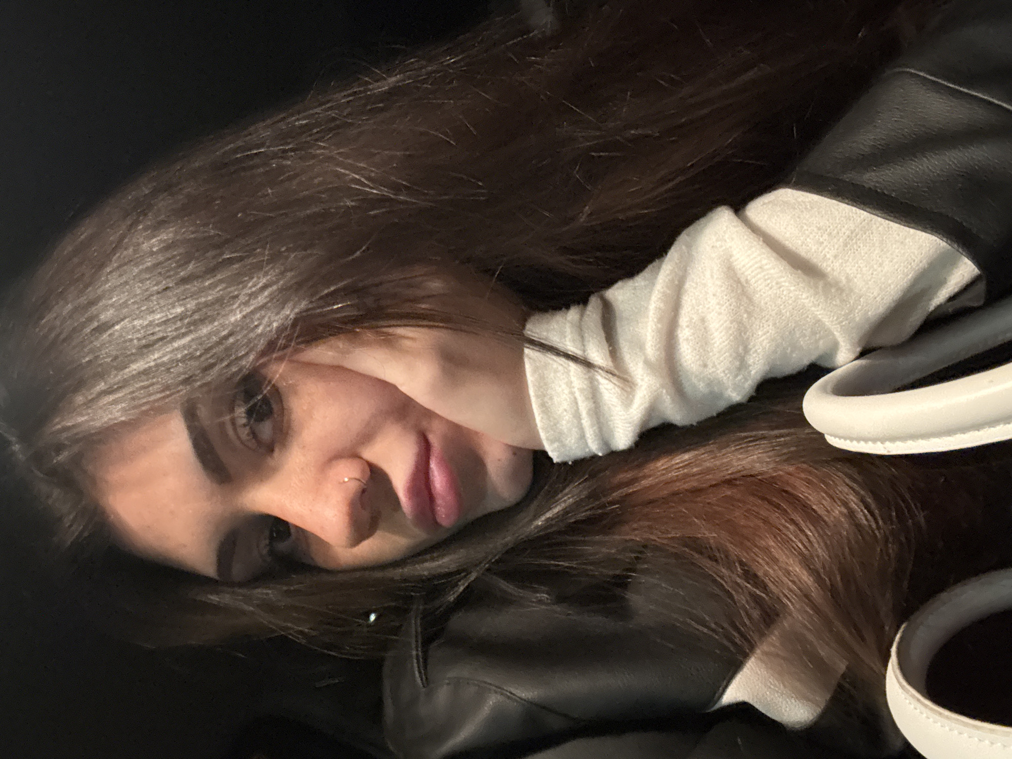 A young woman with long, dark brown hair rests her face on her hand, wearing a white long-sleeved shirt under a black leather jacket. She has a nose piercing on the left side. To the right, a white purse or bag with rounded handles is visible. The background is dark, possibly indicating an indoor setting with low lighting. There is no information to be found that indicates the location of the image in Sofia, Bulgaria.