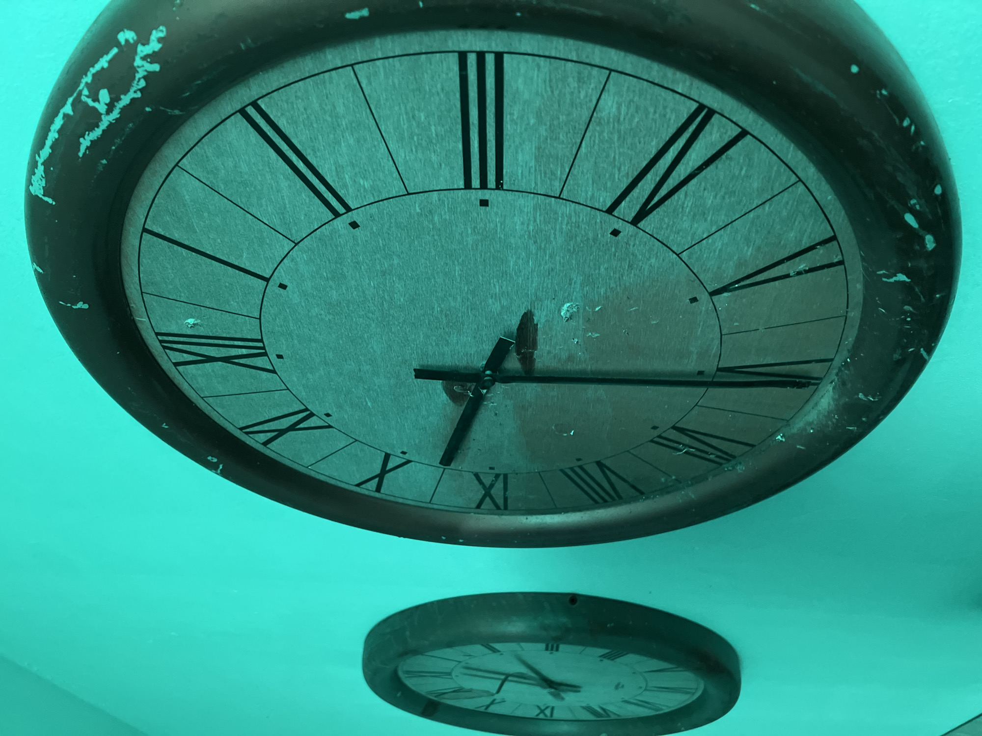 The image displays two wall-mounted analog clocks, both featuring Roman numerals, against a vibrantly colored teal wall. The scene is located in Santeramo in Colle, Italy.
The larger clock occupies the right foreground and is in sharp focus. It is circular with a substantial, dark-colored frame that exhibits significant wear and chipping, particularly noticeable along its upper and lower edges, revealing a lighter material beneath. The clock face is a light teal-grey, displaying black Roman numerals from I to XII. Two black hands are present; the hour hand is positioned slightly past the X, and the minute hand points directly at the IV, indicating a time of approximately 10:20. The central mechanism where the hands pivot shows minor damage or exposure.
A second, smaller circular clock is visible in the left background, out of focus. It possesses a similar dark frame and a light-colored face with dark Roman numerals, consistent in style with the foreground clock.
The background wall is uniformly colored in a vivid teal or aqua hue. It appears to be a plain, unadorned surface, with minor textural variations but no distinct patterns or significant damage. The lighting is diffused, creating subtle shadows beneath both clocks, indicating their protrusion from the wall surface. No individuals or specific actions are depicted within the frame.