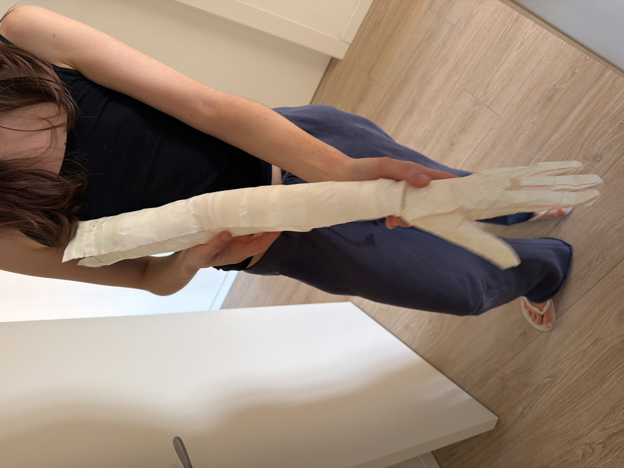 A person is holding a handmade paper hand and forearm cast in their hands, while sitting on a chair in a room with light brown wood flooring. The person is wearing a black tank top and blue sweatpants, and has brown hair. Their feet are clad in white flip-flops. A white table or cabinet is visible along the left edge of the image. The location is not possible to determine from the provided image.
