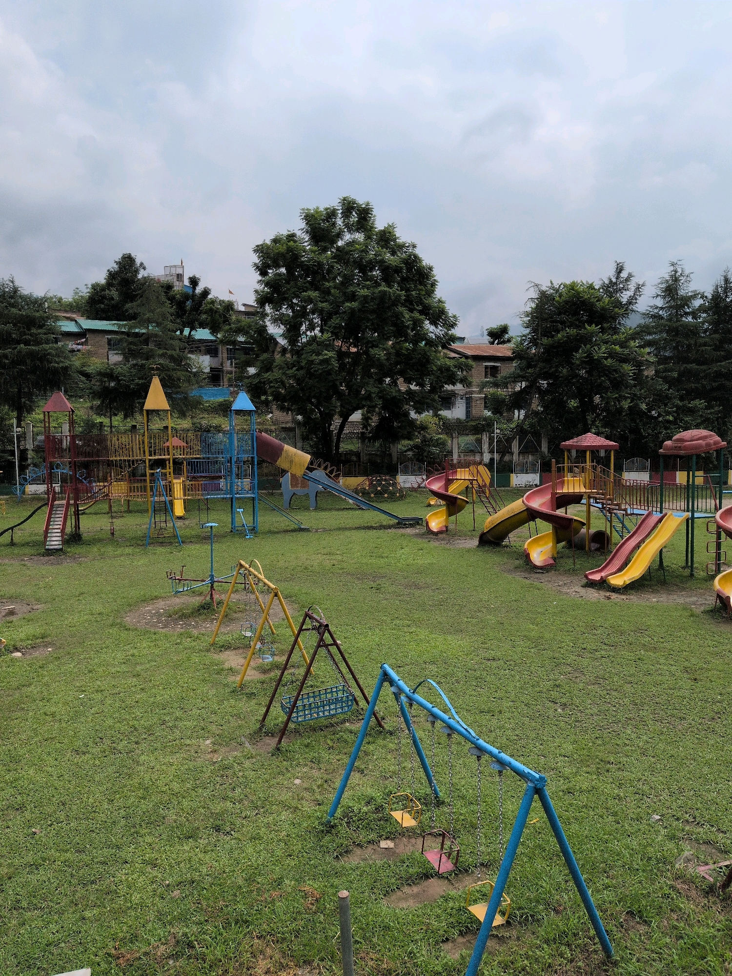 This image captures an empty playground in the city of Mandi, India, on a cloudy day. The playground is situated on a grassy field with several play structures. In the foreground, a blue swing set with two yellow seats and one red seat is visible. Further back, another swing set with yellow supports and blue seats is present. To the left and center, there are various colorful slides, some with yellow and red chutes, integrated into larger play structures with towers and climbing elements. There is also a merry-go-round. In the background, residential buildings with green roofs are nestled among lush green trees, and a pathway is visible. The overall impression is of a quiet, undeveloped public space.