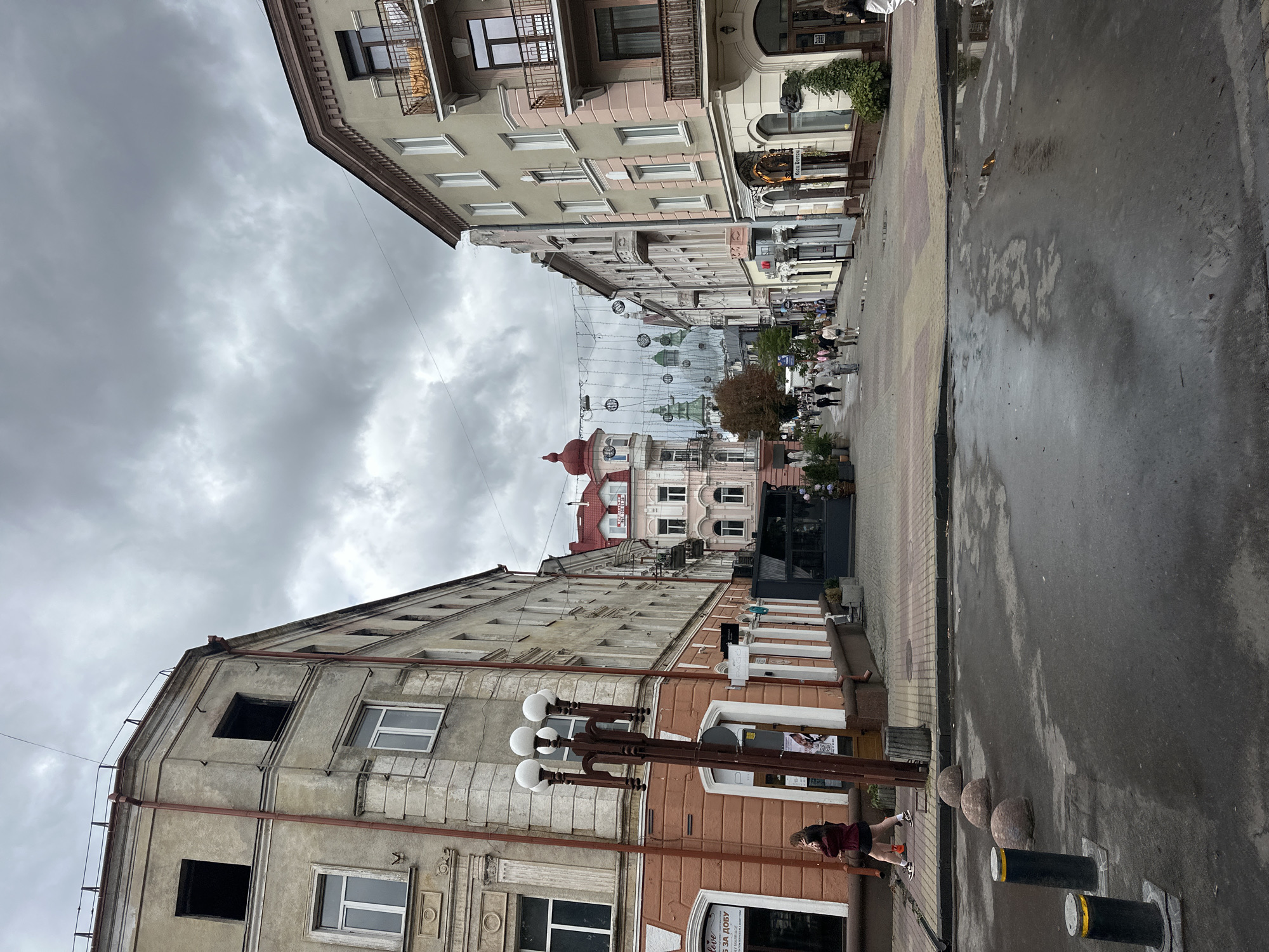 The image depicts an urban street scene in Ternopil, Ukraine, under a cloudy sky. The perspective is from a street-level viewpoint looking down a pedestrianized avenue lined with buildings on both sides.
On the left, a weathered, multi-story building with a salmon-colored lower facade and a pale gray upper facade dominates the foreground. The upper floors show signs of disrepair with dark, empty window openings. A street lamp with five globe-shaped lights stands prominently in front of this building. Two individuals, one in a dark top and skirt, the other in a darker jacket, are walking on the sidewalk.
The right side of the street is also lined with buildings. The closest building on the right has a pale gray and pink facade with multiple windows and balconies. Further down the street, a row of buildings with diverse architectural styles and colors recedes into the distance. Several people are visible walking along the pedestrian area.
Above the street, a decorative string of lights and spherical ornaments is suspended. In the background, several domed structures, possibly churches or historical buildings, are visible. The street surface in the foreground is wet, indicating recent rain, with puddles reflecting the overcast sky and surrounding architecture. Bollards and rounded stone barriers are present along the edge of the street. The overall atmosphere suggests a typical day in a European city, with a mix of old and newer architecture and everyday urban activity.