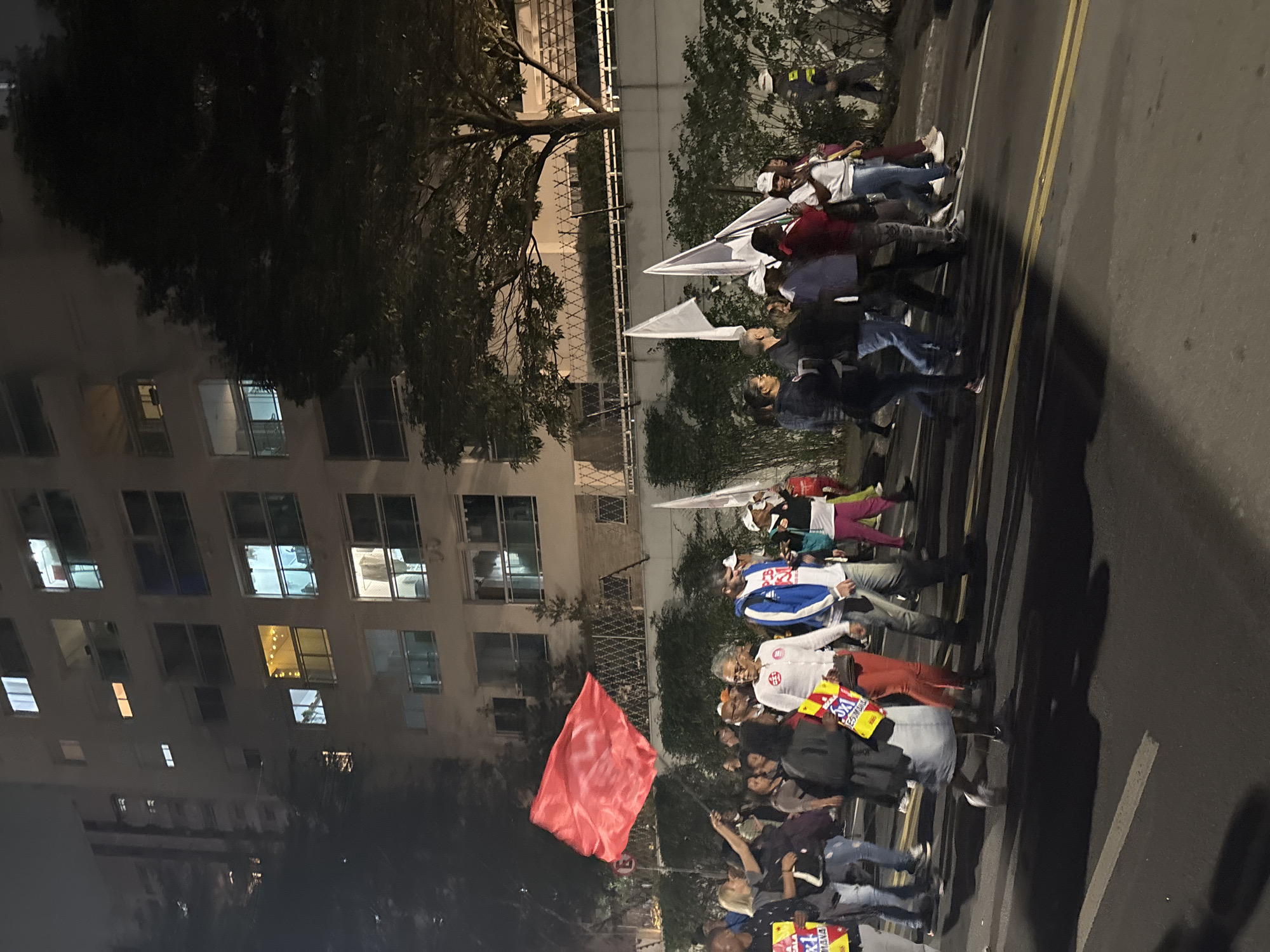 The image depicts a group of people marching along a city street in São Paulo, Brazil. The location is identifiable by the presence of tall buildings with lit windows, a tree-lined street, and a yellow double line on the asphalt. The marchers are carrying various flags and banners, including a red flag and several white flags. They are walking on what appears to be a crosswalk. The time is likely evening or night, given the darkness and illuminated windows. The image is taken from a slightly elevated position, looking down on the march.