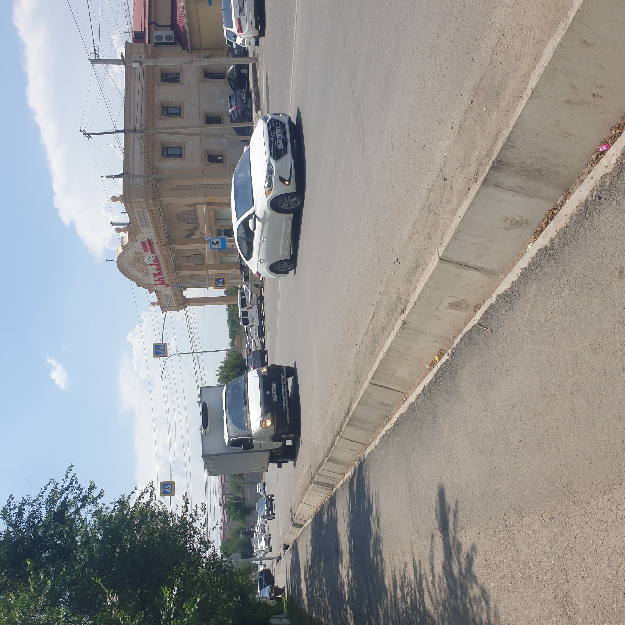 The image captures a street scene in Shymkent, Kazakhstan, on a sunny day with scattered clouds. A white truck is positioned on the right side of the road, facing left, with its headlights on. Further down the road, a white Hyundai Elantra is driving towards the viewer, also with its headlights illuminated. On the left side of the road, a sidewalk is visible, shaded by trees. Beyond the sidewalk, several buildings are present, with one prominent building featuring an ornate facade with the inscription "Jubilee" in red lettering. Road signs, including a pedestrian crossing sign and a fuel pump sign, are visible. Power lines crisscross the sky above the street. The road surface is asphalt, with a concrete curb separating the road from the sidewalk.