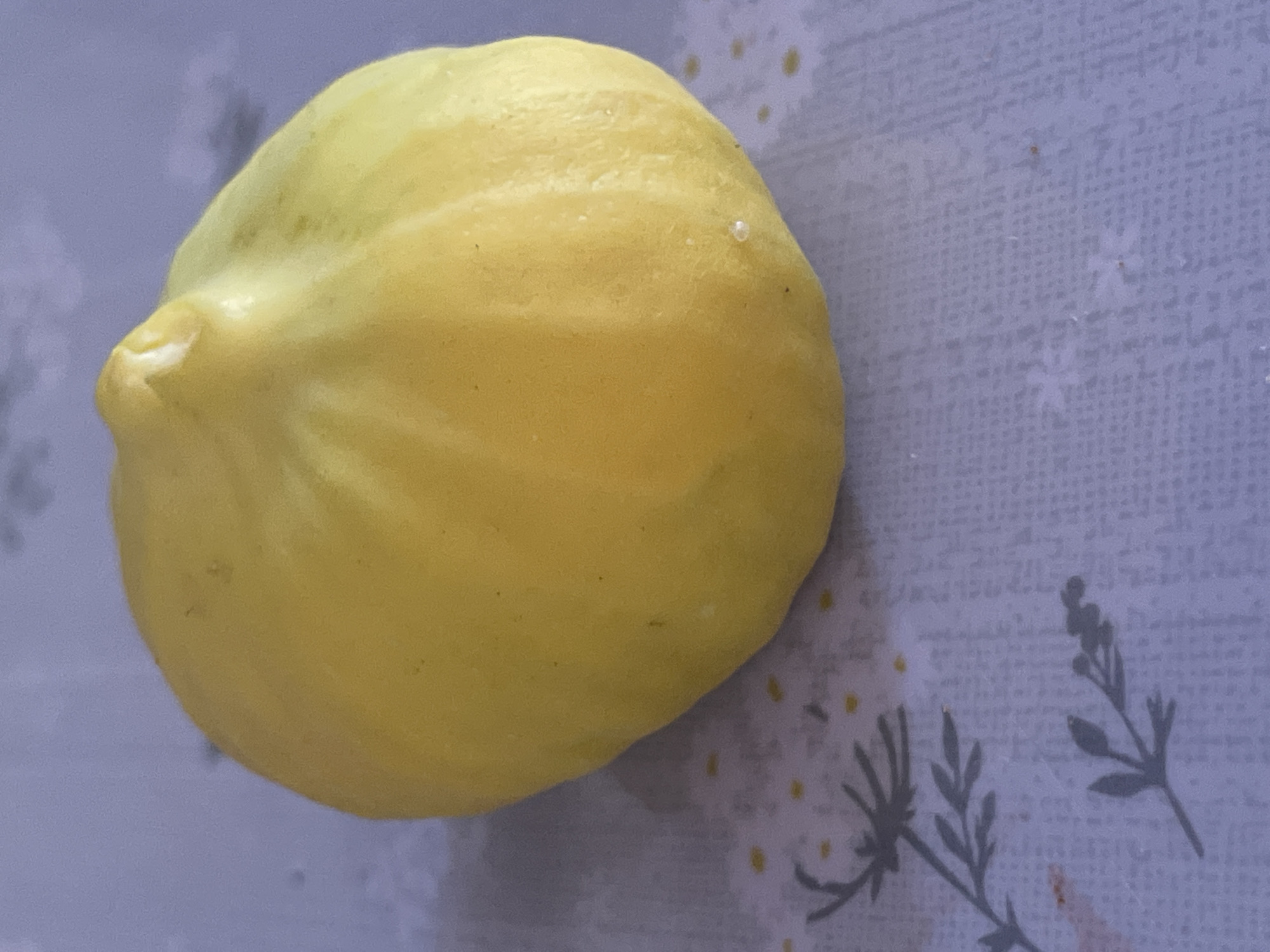 The image shows a single, ripe, yellow fig placed on a light-colored patterned surface. The surface appears to be a tablecloth or placemat with a subtle floral design and scattered small yellow dots. The fig is oval-shaped with a slightly irregular top, exhibiting subtle ridges and color variations, with some areas appearing lighter yellow or greenish. The background is out of focus and appears to be a solid, muted color. There is no indication of a city or specific location within Tasbuget, Kazakhstan, in this image.
