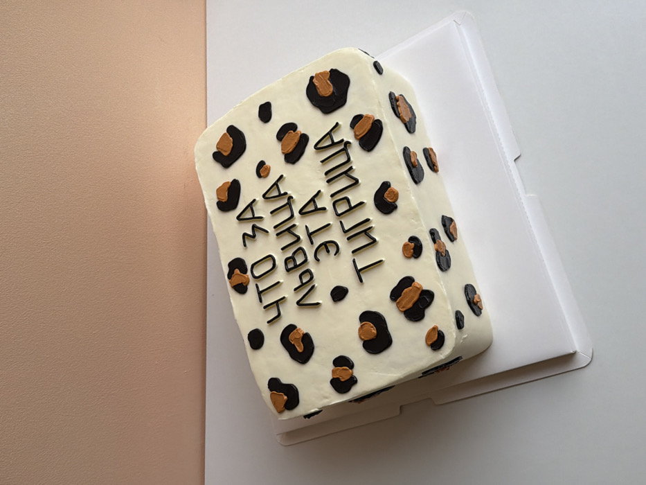 A rectangular cake sits atop a white cardboard base against a tan wall. The cake is covered in white frosting decorated with black and gold leopard spots. Black text in a serif font is written on the side of the cake. The cake appears to be homemade, as the frosting is not perfectly smooth and the leopard spots are slightly asymmetrical.
