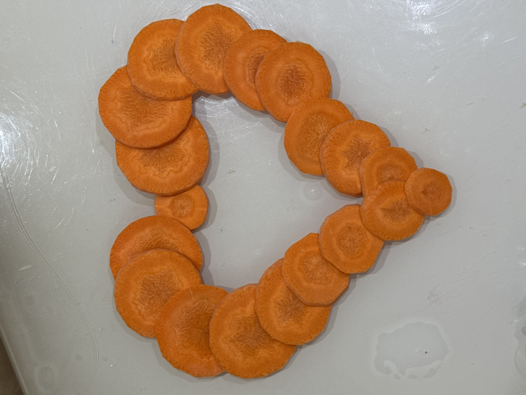 The image shows carrot slices arranged in the shape of a heart on a light-colored, possibly plastic, surface. The carrot slices are round and orange, with visible internal texture. The lighting appears to be from above, casting slight shadows. The surface has some scuff marks and a small water stain. There are no people or buildings visible. This image is not related to the city of Salsk, Russia.