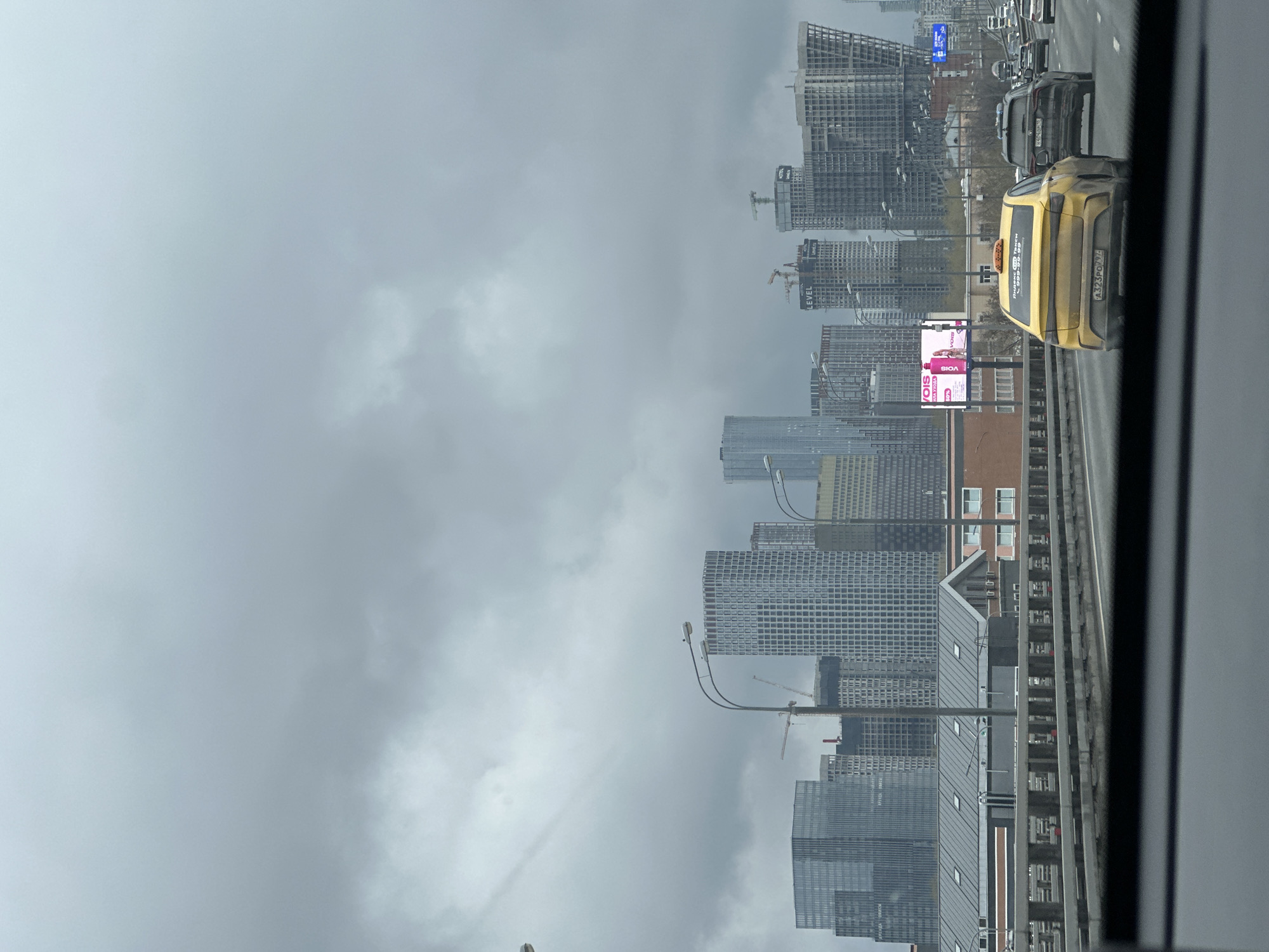 The image shows a view from inside a vehicle, likely on a highway, overlooking the cityscape of Moscow, Russia. The sky is overcast with grey clouds. Several modern high-rise buildings are visible, many of which appear to be under construction with cranes. In the foreground, there are multiple vehicles on the highway, including a yellow taxi with Russian license plates. A billboard with a pink advertisement is also visible. The road appears to be elevated.