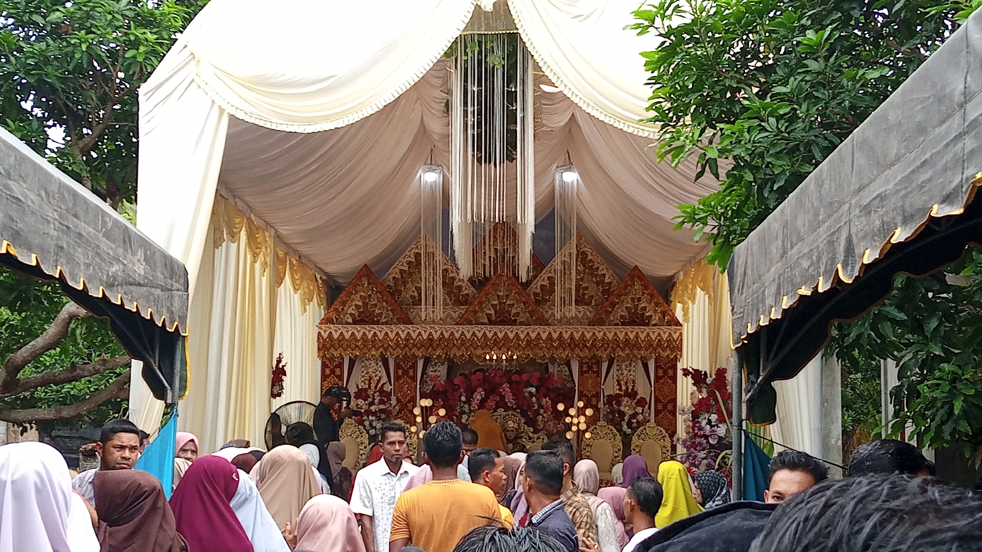 This image depicts an outdoor wedding ceremony in Arabungong, Indonesia. The scene is dominated by a large, ornate white tent with gold trim, under which a wedding stage is set up. The stage features a traditional Indonesian wedding altar decorated with red and pink flowers, gold accents, and intricate textile patterns. Several guests, mostly women wearing hijabs in various colors, are gathered around the stage, observing the ceremony. Some men are also present, some in the foreground looking towards the stage, others in the background near the altar. Lush green trees and foliage frame the scene, suggesting a natural or garden setting. The overall atmosphere appears festive and ceremonial.