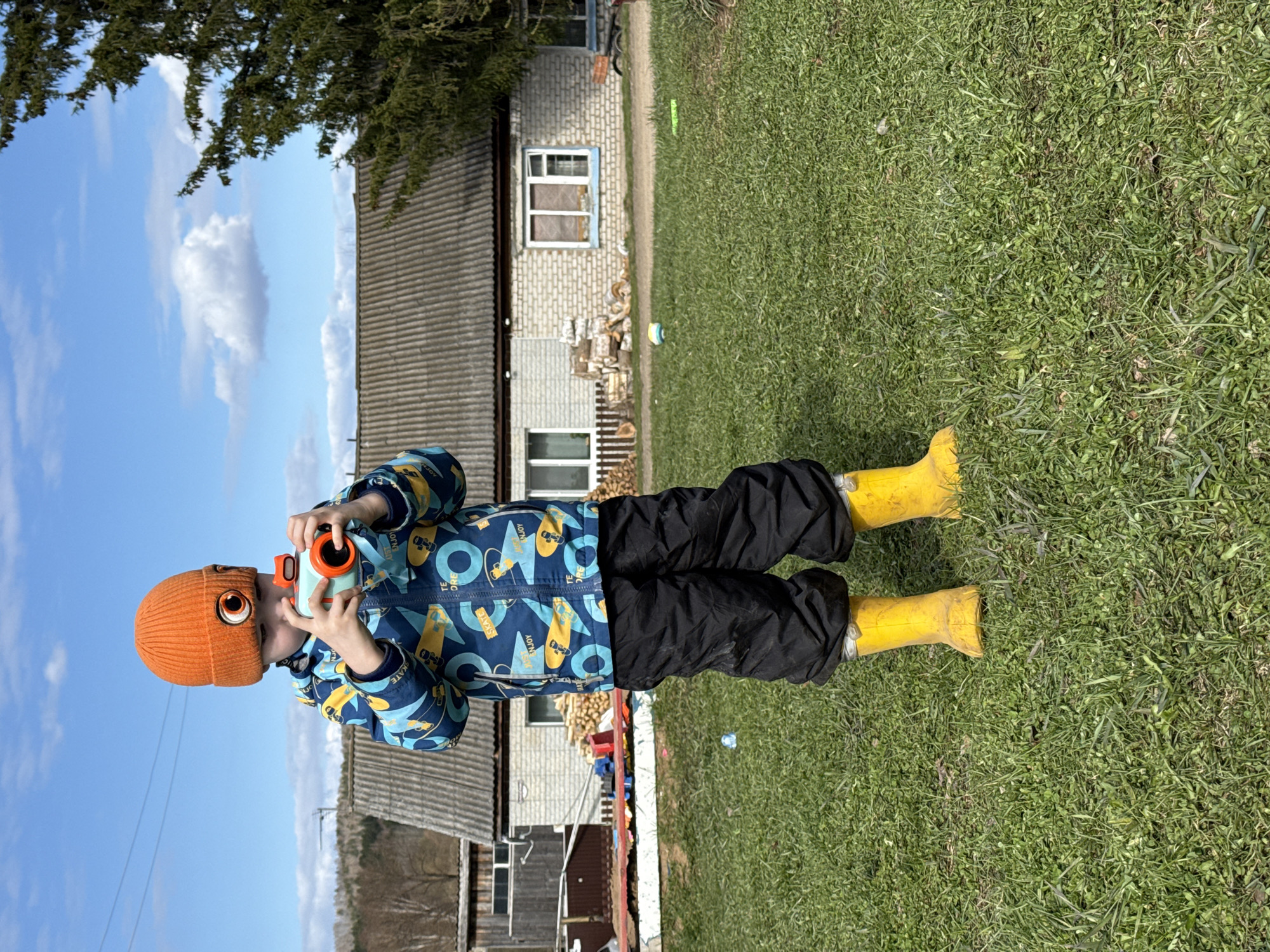 A young child wearing an orange knit hat, a blue and yellow patterned jacket, black pants, and yellow boots stands on a grassy lawn holding a toy camera. The child is positioned facing toward the left of the image and is centered. Behind the child is a light-colored brick building with a grey roof and white-framed windows. Stacks of wood are visible near the building. A blue sky with scattered clouds is visible in the upper left corner. The environment suggests a rural or suburban setting, potentially in Nizhnyaya Chermoda, Russia.