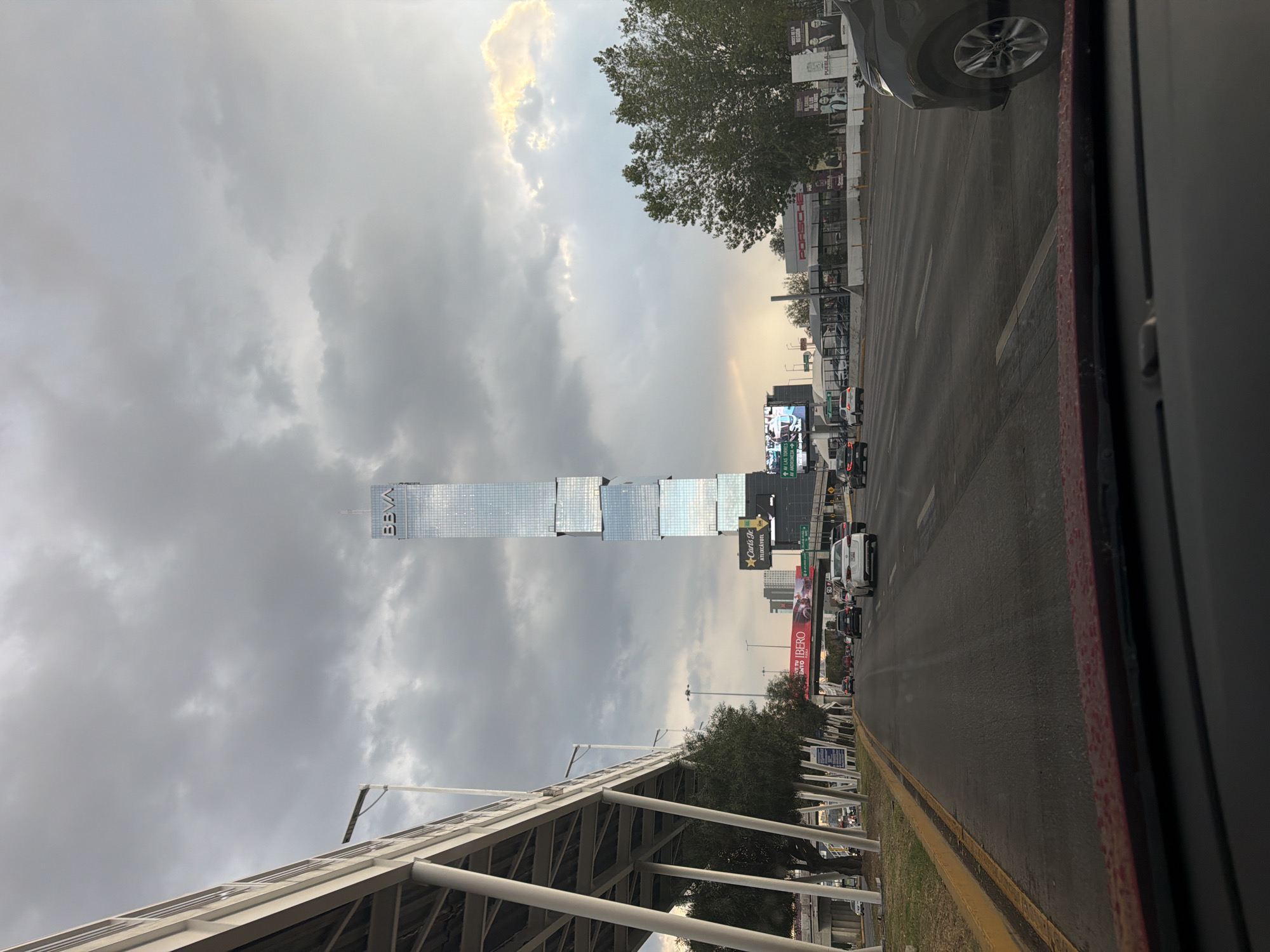 The image depicts a street scene in Heroica Puebla de Zaragoza, Mexico. A tall, modern skyscraper with the "BBVA" logo is visible against a cloudy sky. A multi-lane road with cars in traffic runs in front of the building. A pedestrian bridge is located on the lower left side of the image. Trees and various billboards line the street. The photograph appears to have been taken from inside a vehicle.