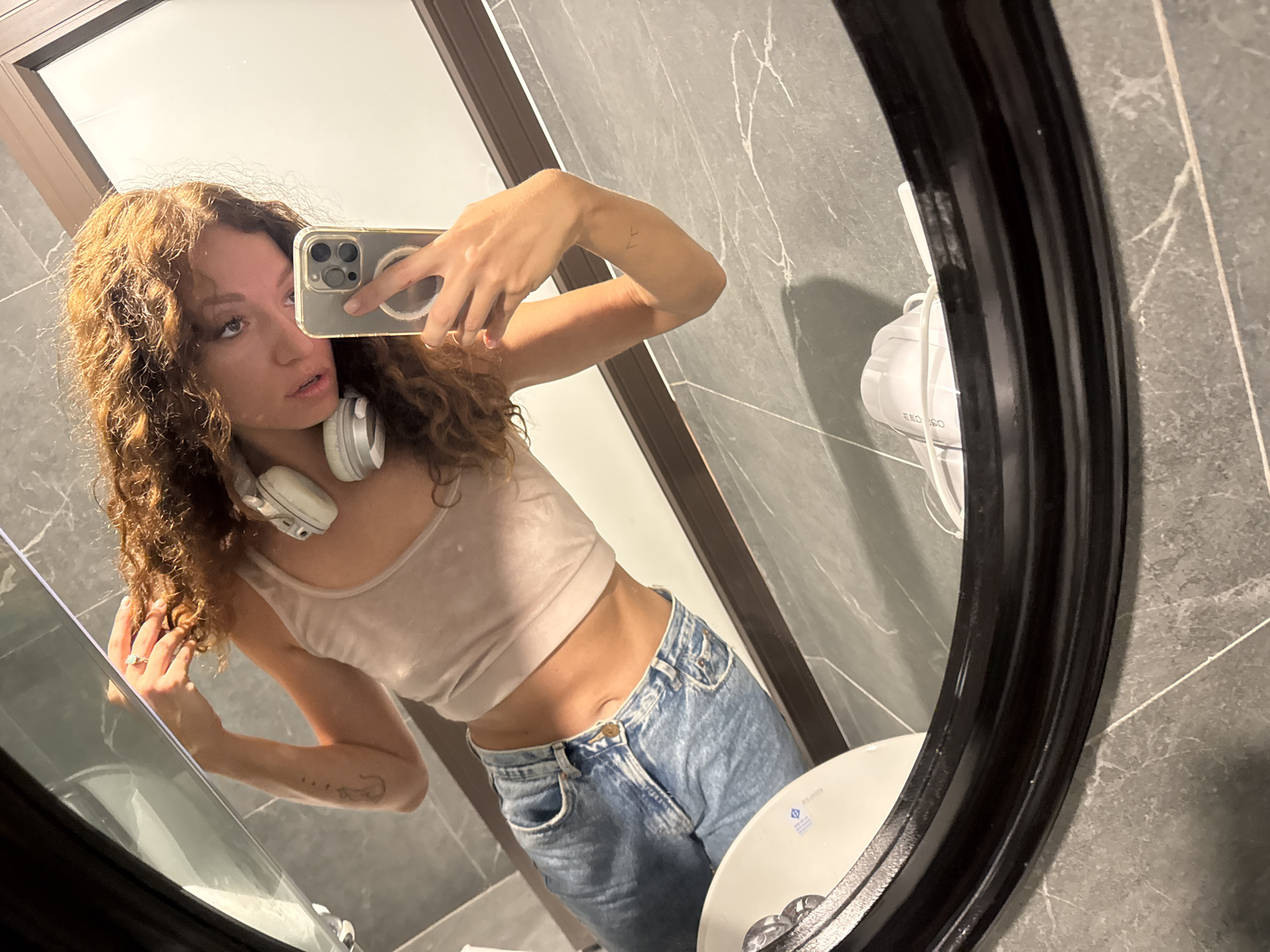 A young woman is taking a selfie in a bathroom. She is wearing a light-colored tank top and light blue jeans. Headphones are around her neck. The phone is in her right hand. There is a mirror that is oval-shaped with a dark frame. The bathroom has grey marble tiles. A hair dryer is mounted on the wall next to the mirror. The city of Hà Nội, Vietnam is not discernible in the image.