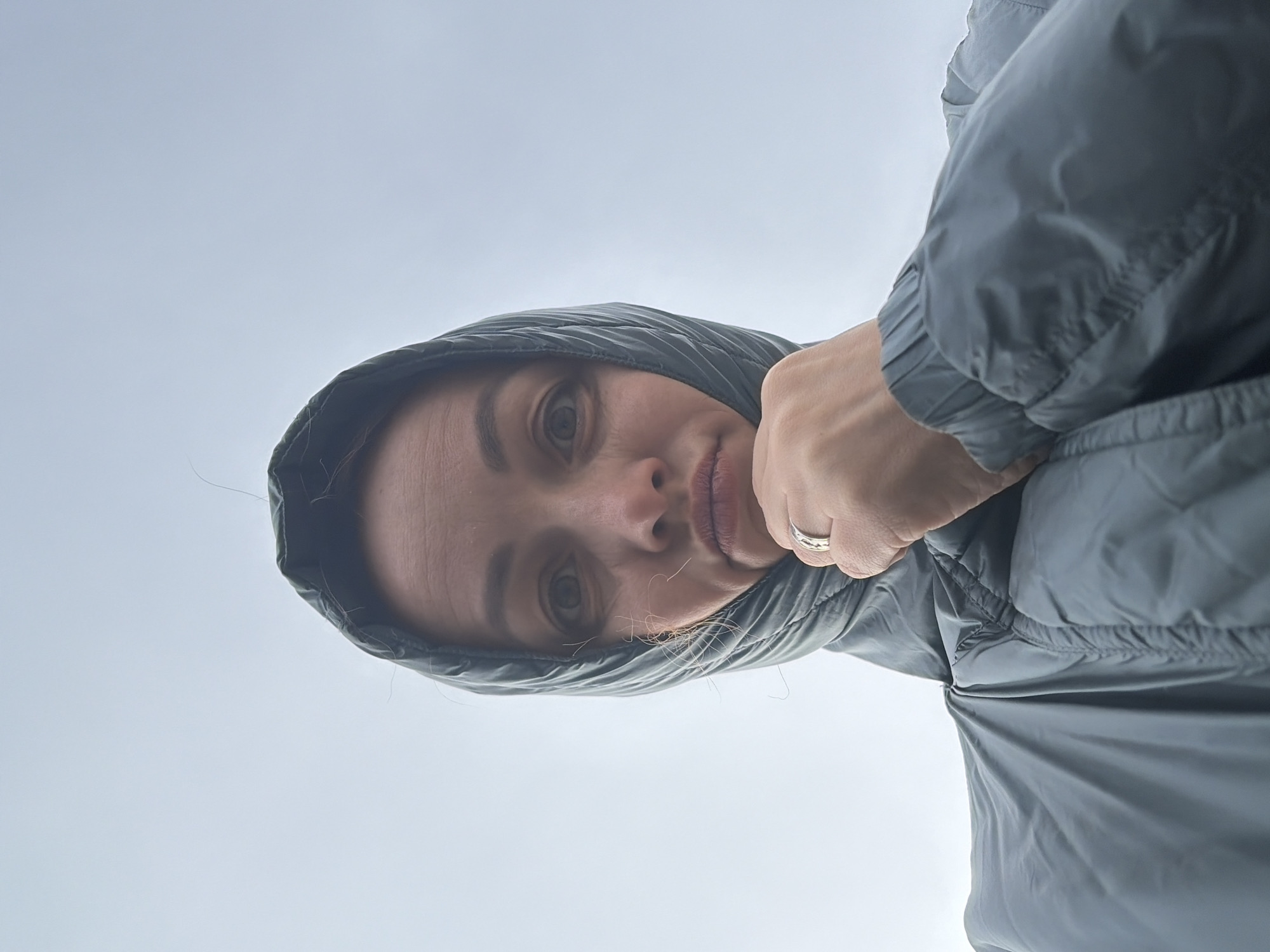 A woman in a grey hooded jacket looks down at the camera from a low angle. She has light skin, light eyes, and thin eyebrows. Her lips are slightly parted. She is wearing a wedding ring on her left hand. The background is a cloudy, light grey sky. The image was not taken in Chaikovsky, Russia.