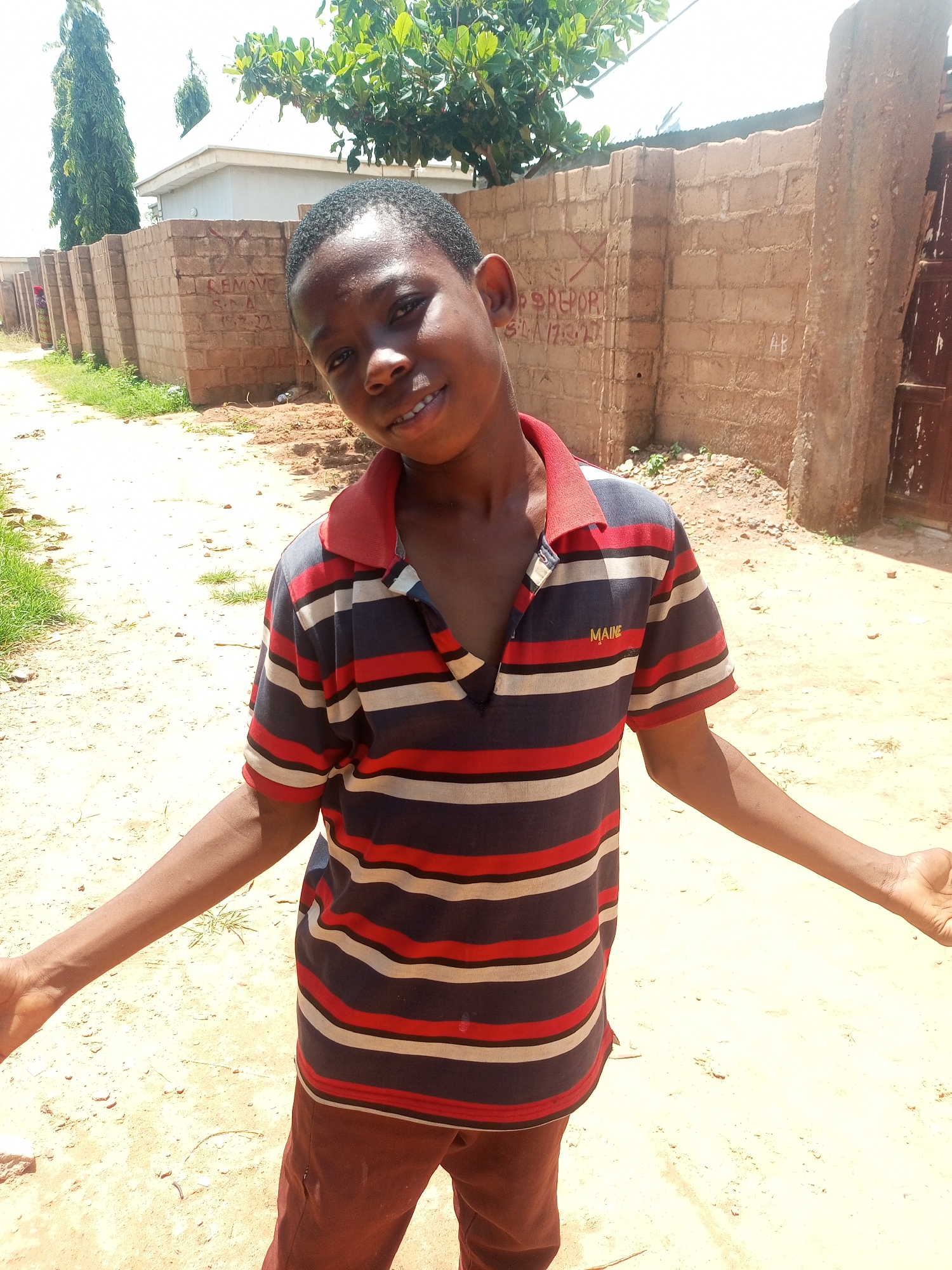 In the city of Tempure, Nigeria, a young boy stands outdoors in front of a brick wall. He is wearing a horizontally striped polo shirt with red, white, and dark blue bands, and dark red pants. His arms are extended to his sides, and he is smiling at the camera. The ground is covered in sand and scattered debris. Behind him, the brick wall is visible, with some graffiti on it. Trees and part of a building are also in the background. The sky is bright, indicating a sunny day.