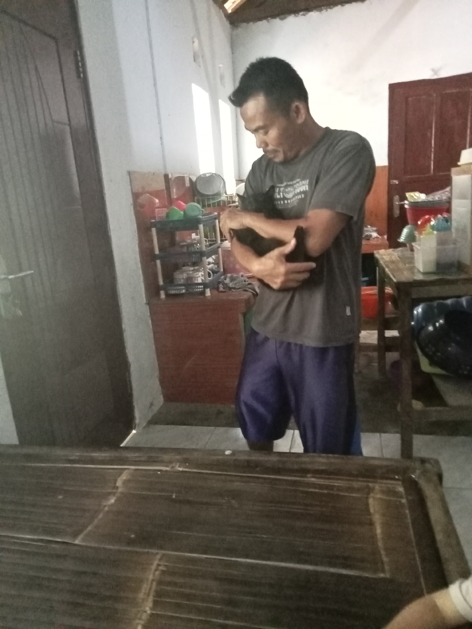 A man in a gray t-shirt and blue shorts is holding a black cat. He is standing in a room with a tiled floor and a wooden table in the foreground. There is a shelf unit with various items on it and a red door in the background. The lighting suggests it is daytime. The scene appears to be indoors, possibly a residence or small shop in Panyaungan Jaya, Indonesia.