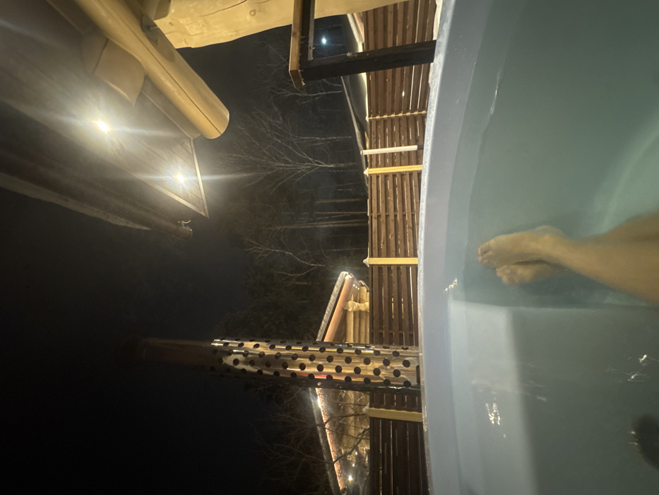 The image depicts an outdoor night scene in Krasnoyarsk, Russia, featuring a person's bare feet and lower legs submerged in the light blue water of a hot tub, viewed from a first-person perspective. The water surface shows slight ripples.
Immediately beyond the hot tub, a dark wooden slatted fence extends horizontally across the midground. To the left, a tall, cylindrical metal chimney or heating element with a patterned perforation of circular holes rises from behind the fence, emitting an orange glow from its interior base. Further to the left and behind the fence, a log cabin-style building with an illuminated roofline is partially visible.
The background consists of a dark night sky above a dense stand of bare, deciduous trees. In the upper right quadrant, a robust wooden beam structure, possibly an overhang or roofline, is present, featuring two bright, recessed lights that cast distinct light rays downwards. A large, light-colored wooden log is also partially visible on the far right, forming part of the overhead structure. The scene is artificially lit, suggesting a recreational or leisure setting in a cold climate.
