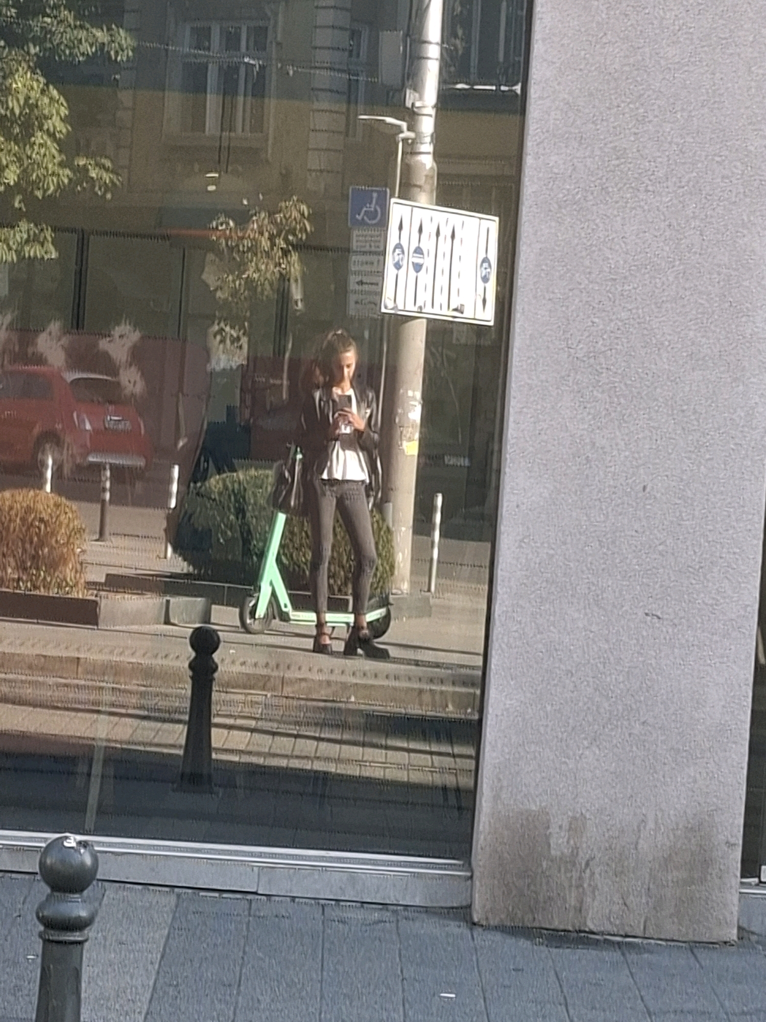 In an urban setting, a young woman stands on a sidewalk, holding a smartphone and looking down at it. She is dressed in a white top, dark jeans, and a dark jacket, and she is wearing black shoes. To her left, a green electric scooter is parked. Behind her, a red car is visible, and further in the background, a building with windows can be seen. A traffic sign with multiple arrows is mounted on a pole to the left of the woman. The scene is reflected in a large glass window, creating a mirrored image of the surroundings. A grey concrete pillar is on the right side of the frame.