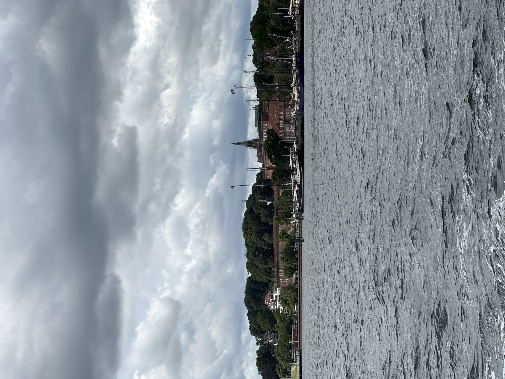 This image shows a cloudy sky over the harbor of Kiel, Germany, with a view of the city's waterfront. In the foreground, dark, choppy water dominates the lower half of the frame, with whitecaps visible. The middle ground features a shoreline lined with trees and buildings. Several sailboats and larger vessels are docked along the waterfront. Behind the boats, a cluster of brick buildings, some with red tile roofs, can be seen. One building has a distinctive spire. The sky is filled with heavy, gray, overcast clouds, suggesting inclement weather.