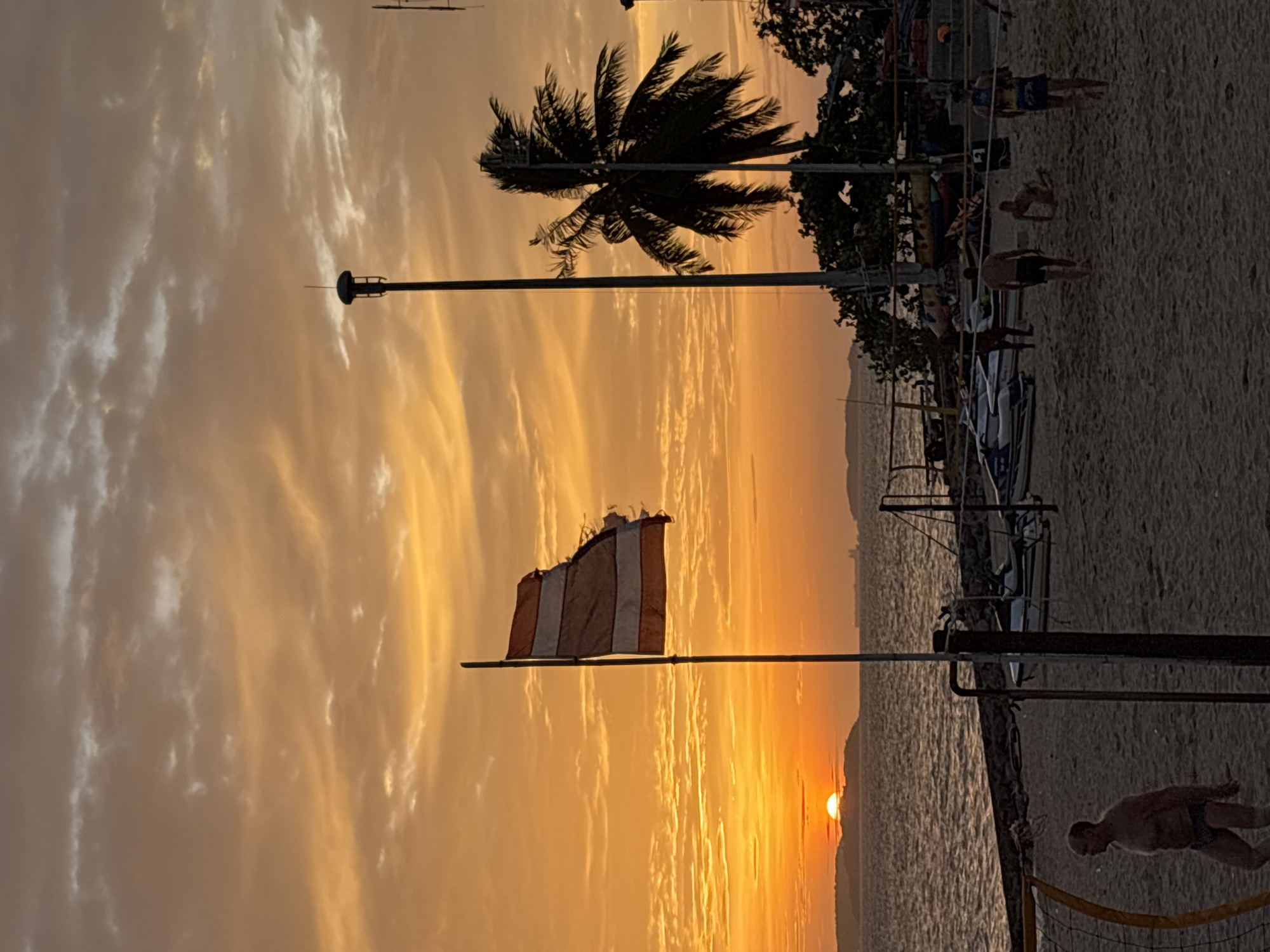 The image depicts a sunset scene on a beach in Pattaya City, Thailand. The sky is filled with vibrant orange and yellow hues, with scattered clouds. In the foreground, a beach volleyball net is partially visible. Several people are present on the sandy beach; some are walking, and others appear to be playing or observing the sunset. A man in swim trunks is walking on the left side of the frame. In the mid-ground, there are several jet skis parked on the beach and in the water. A palm tree stands tall on the right side of the image. A tattered flag, possibly a Thai flag, is flying on a pole in the left-center of the frame. The ocean is visible in the background, with a faint outline of land or islands on the horizon. The overall atmosphere is that of a relaxed evening at a popular tourist destination.
