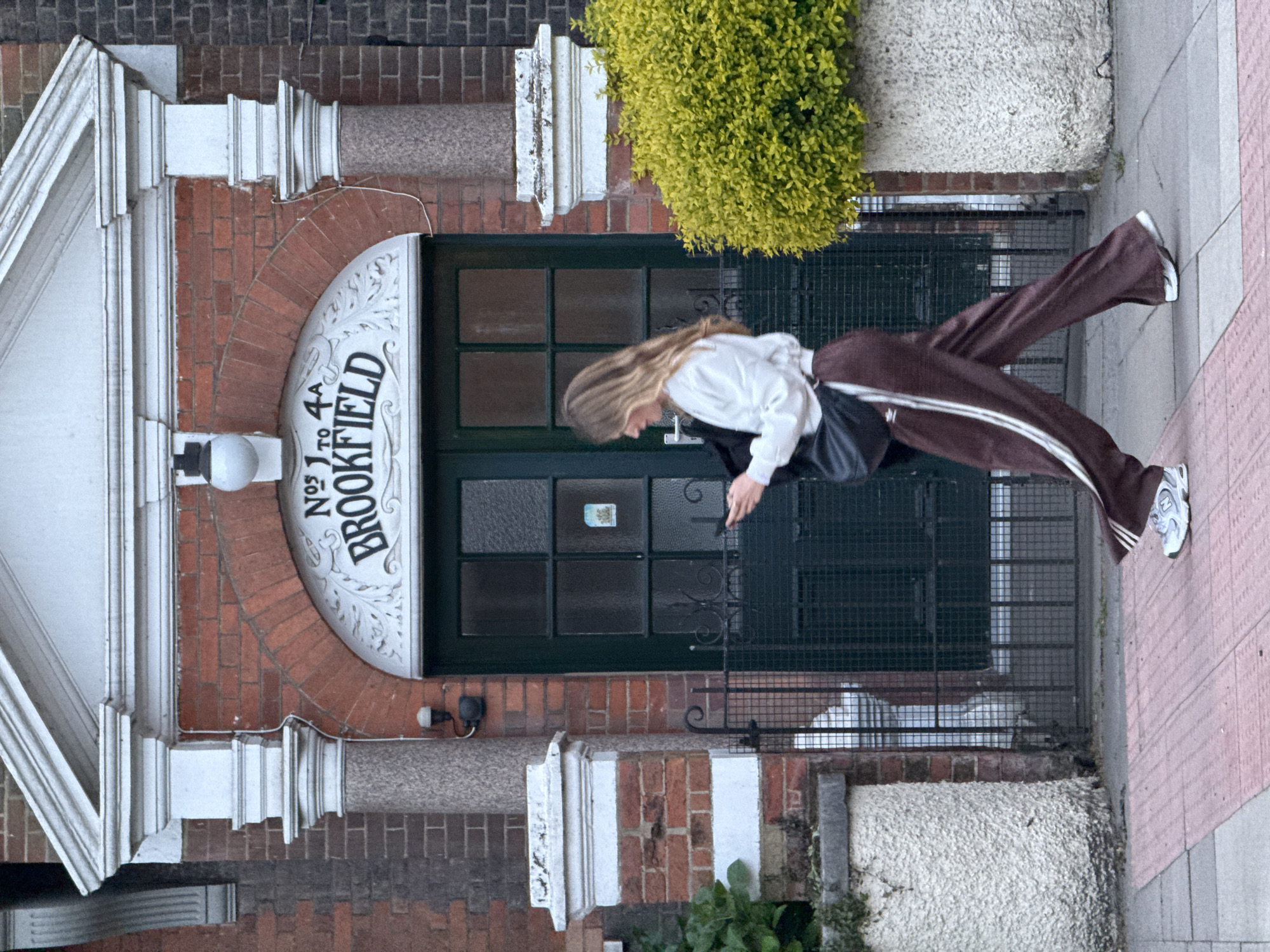 A person with long blonde hair wearing a white top and brown track pants with white stripes is standing in front of a building with the inscription "Nos 1 to 4A Brookfield." The building is made of red brick with white architectural details and a dark green door with window panes. A leafy bush is situated to the right of the entrance. The person is looking down, possibly at a device in their hands, and is standing on a sidewalk paved with pink and grey slabs. The location appears to be a building entrance in the city of London, United Kingdom.