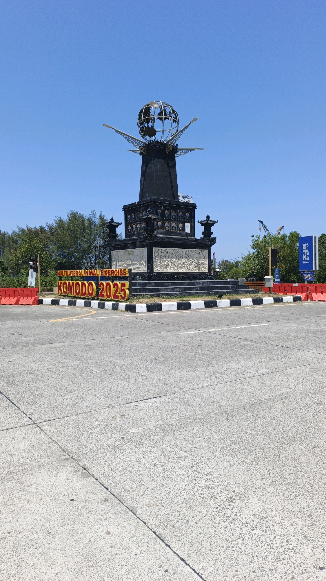 In Denpasar, Indonesia, a monument stands tall against a clear blue sky. The monument is a large, dark structure topped with a globe supported by wings. Below the globe, intricate carvings adorn the monument's base. In front of the monument, a sign reads "MULTILATERAL NAVAL EXERCISE KOMODO 2025" in large, bright letters. The monument is situated at an intersection with a paved road. Red and white traffic barriers are visible on the left and right sides of the monument, and some trees are in the background. There are no people visible in the image.