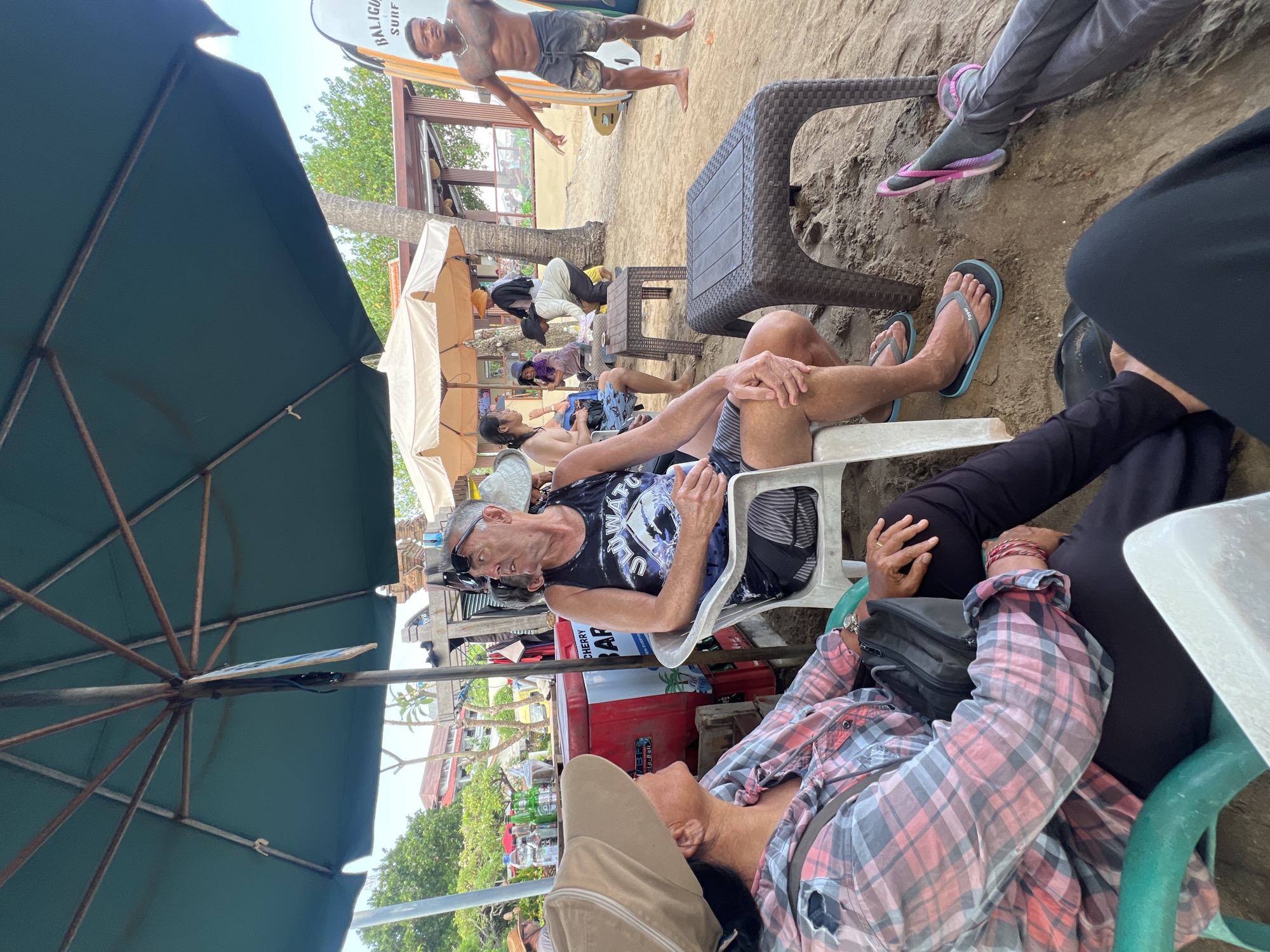 This image depicts a casual scene on a beach in Kuta, Indonesia. In the foreground, a woman wearing a wide-brimmed hat and a pink and grey plaid shirt is seated in a green chair, facing away from the camera. Next to her, a man in a black tank top and shorts is seated in a white chair, looking to his right. Both are under a large green umbrella. Behind them, other individuals are visible, some sitting and others standing. One man, shirtless and wearing dark shorts, stands near several surfboards, gesturing with his right hand. In the background, a red cooler with "CHERRY BAR" written on it is visible, along with assorted bottles. The environment is sandy, with palm trees and beach structures suggesting a commercialized beach area. The lighting indicates it is daytime.