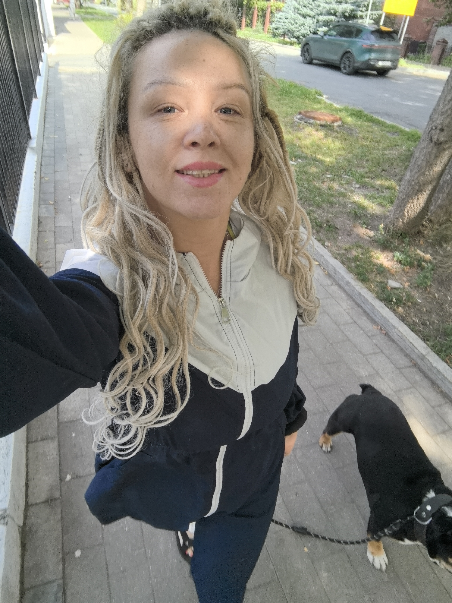 A woman with blonde dreadlocks is walking her black and tan dog on a leash on a paved sidewalk. She is wearing a dark blue and white jacket and dark pants. The dog is walking ahead of her, and its leash is attached to her hand. The sidewalk is next to a grassy area with a tree. In the background, a green car is driving on a street. The scene is taking place during the day. The location is Nizhny Novgorod, Russia.