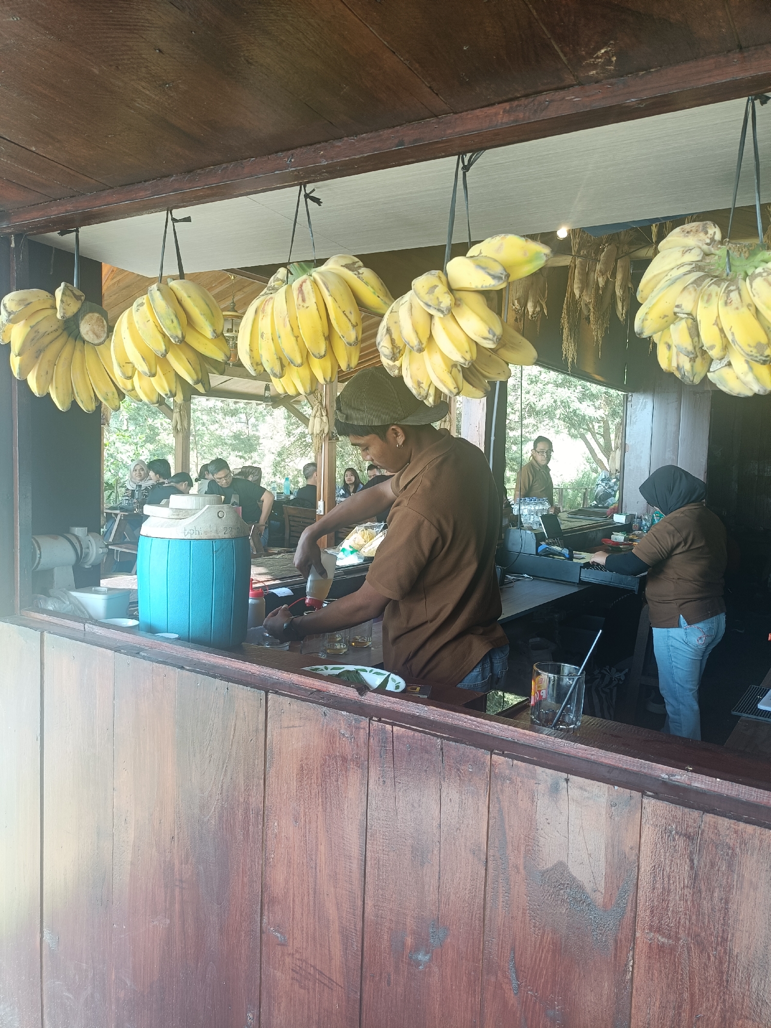 In Kopeng, Indonesia, a rustic wooden counter is the setting for a scene of beverage service. Bunches of ripe yellow bananas hang from the ceiling, a tropical motif evident in the open-air establishment. A man, wearing a brown short-sleeved shirt and a cap, is behind the counter, preparing a drink. He is pouring a liquid from a bottle into a glass. Next to him, a large blue cooler sits prominently. Further down the counter, a woman in a brown long-sleeved shirt and a hijab, wearing blue jeans, operates a cash register. In the background, patrons are seated at tables, enjoying the ambiance. The overall impression is that of a casual, outdoor cafe or eatery.