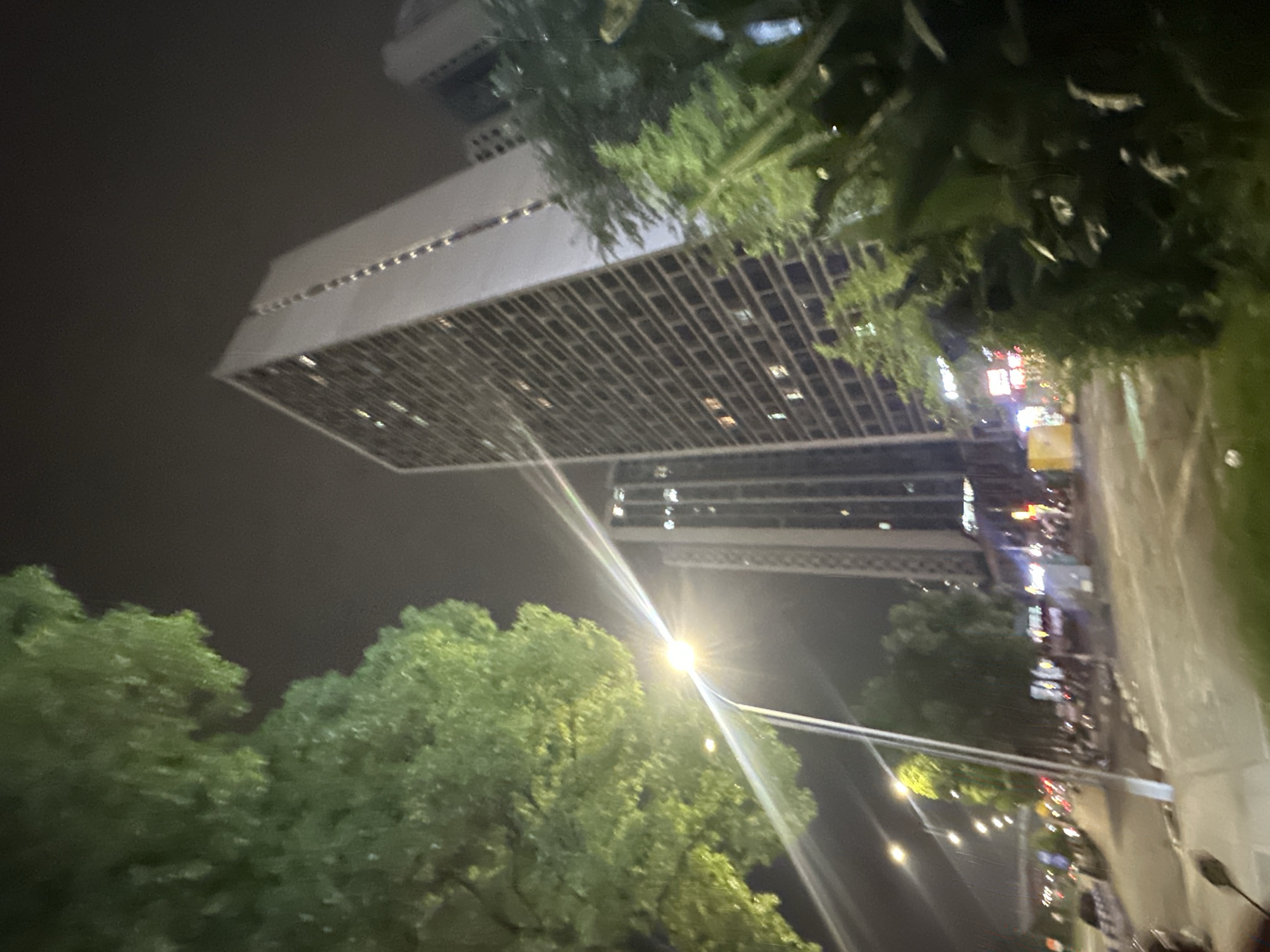 The image depicts a nighttime street scene in the Chongqing High-tech Zone, China. Tall buildings dominate the background, with one particularly prominent, multi-story structure displaying numerous lit windows. In the foreground, a large tree with green foliage partially obscures the view. A street lamp emits a bright light, creating a lens flare effect that streaks across the image. The street itself appears to be a road with traffic, indicated by the presence of vehicles and streetlights. Some of the buildings have illuminated signs, suggesting commercial activity. The overall atmosphere is urban and active, even at night.