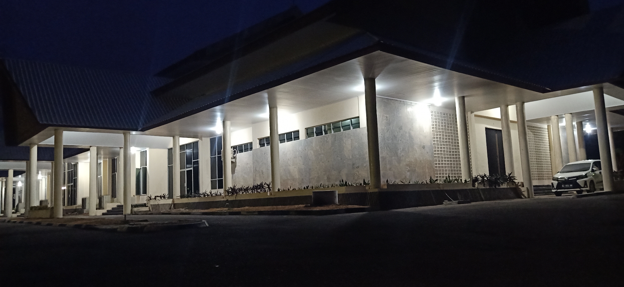 The image shows the exterior of a building at night, located in Palohgadeng, Indonesia. The building features a modern architectural style with a large, covered walkway supported by numerous columns. The walkway is illuminated by overhead lights, casting a warm glow on the polished concrete floor and the beige-colored walls. Large windows with dark frames are visible along the building's facade, some reflecting the internal lights. A white car is parked to the right of the building, its headlights off. The ground in front of the building is paved with asphalt, and there are some low-lying plants and planters along the edge of the walkway. The sky above is dark blue, indicating nighttime.