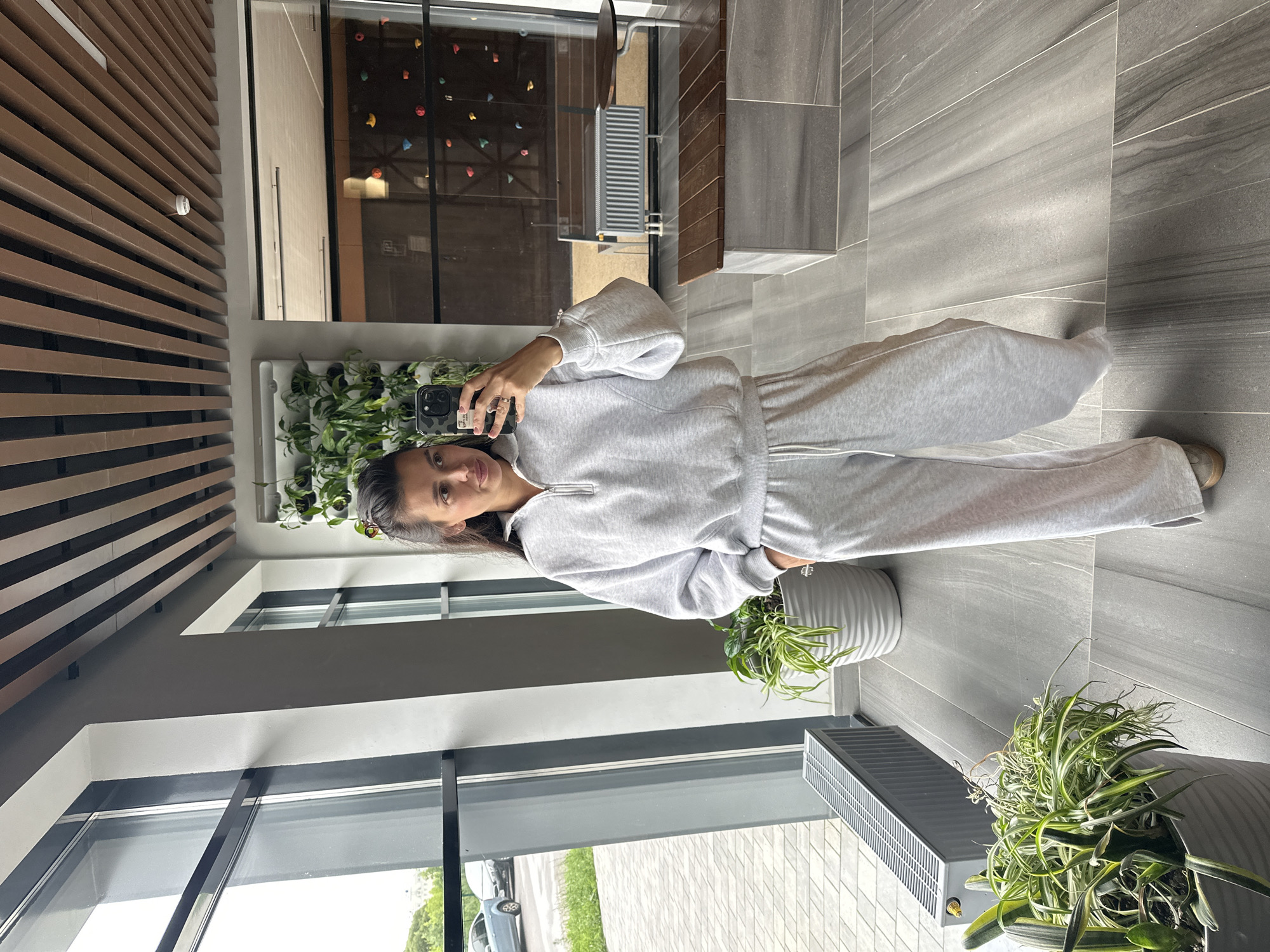 A woman is taking a mirror selfie in an indoor setting. She is wearing a light gray loungewear set consisting of a quarter-zip sweatshirt and wide-leg sweatpants. The background appears to be a modern lobby or common area with large windows, a vertical garden on the wall, and a climbing wall visible through a glass partition. There are plants in pots and a radiator unit present. The flooring is made of large gray tiles. The woman's posture suggests she is standing casually. The overall environment is clean and contemporary. The location is identified as being in the city of Perm, Russia.
