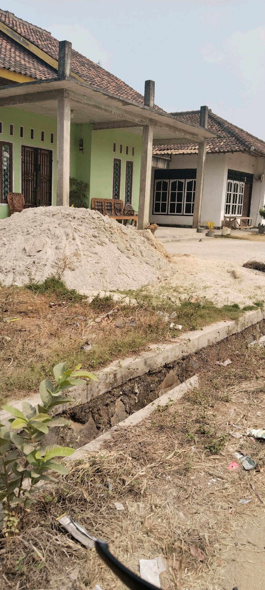 The image depicts a street scene in Tanjung Ratu Ilir, Indonesia, featuring two adjacent residential buildings. The building on the left is painted light green and has a covered porch supported by concrete columns. A large pile of sand is located in front of this building, obscuring part of the lower facade. The building on the right is white and also has a covered porch. Both buildings have tiled roofs and visible windows. The area in front of the buildings appears to be a pathway or driveway, with some vegetation and dry grass growing around it. A concrete drainage ditch runs along the foreground of the image, filled with debris and stagnant water. The overall scene suggests a residential area undergoing construction or renovation, with elements of disarray and ongoing work.