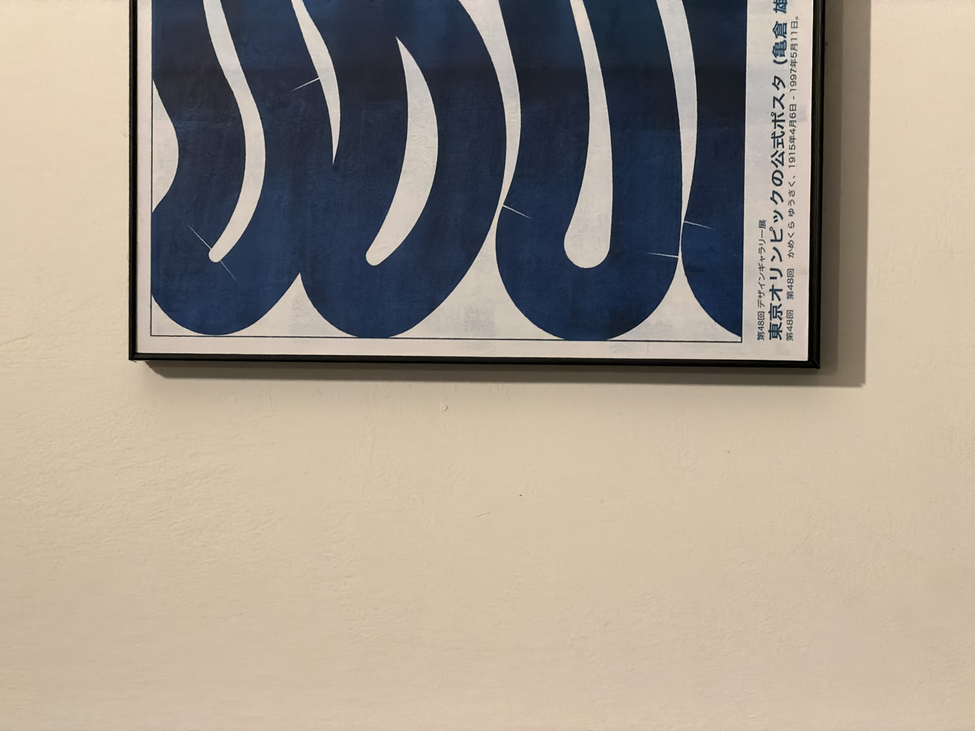 A framed poster is depicted hanging on a light-colored, textured wall in an indoor setting. The poster features a large, abstract design dominated by sweeping, wave-like or calligraphic forms in deep blue against a white background. The design is dynamic, with the dark blue shapes appearing to intertwine and flow across the frame. The poster is encased in a slim black frame.
On the right side of the poster, vertically aligned Japanese text provides details about the artwork. The text identifies it as the "48th Design Gallery Exhibition" and the "Official Poster for the Tokyo Olympics," attributed to "Yusaku Kamekura." It also includes Kamekura's birth and death dates: "April 6, 1915 - May 11, 1997."
The wall itself is a plain, neutral tone, possibly off-white or light beige, showing subtle texture. There are no people or discernible activities taking place, and the scene is static. The lighting is even, typical of an indoor environment. While the location context is given as North Kuta, Indonesia, no specific visual cues within the image confirm this geographical setting.