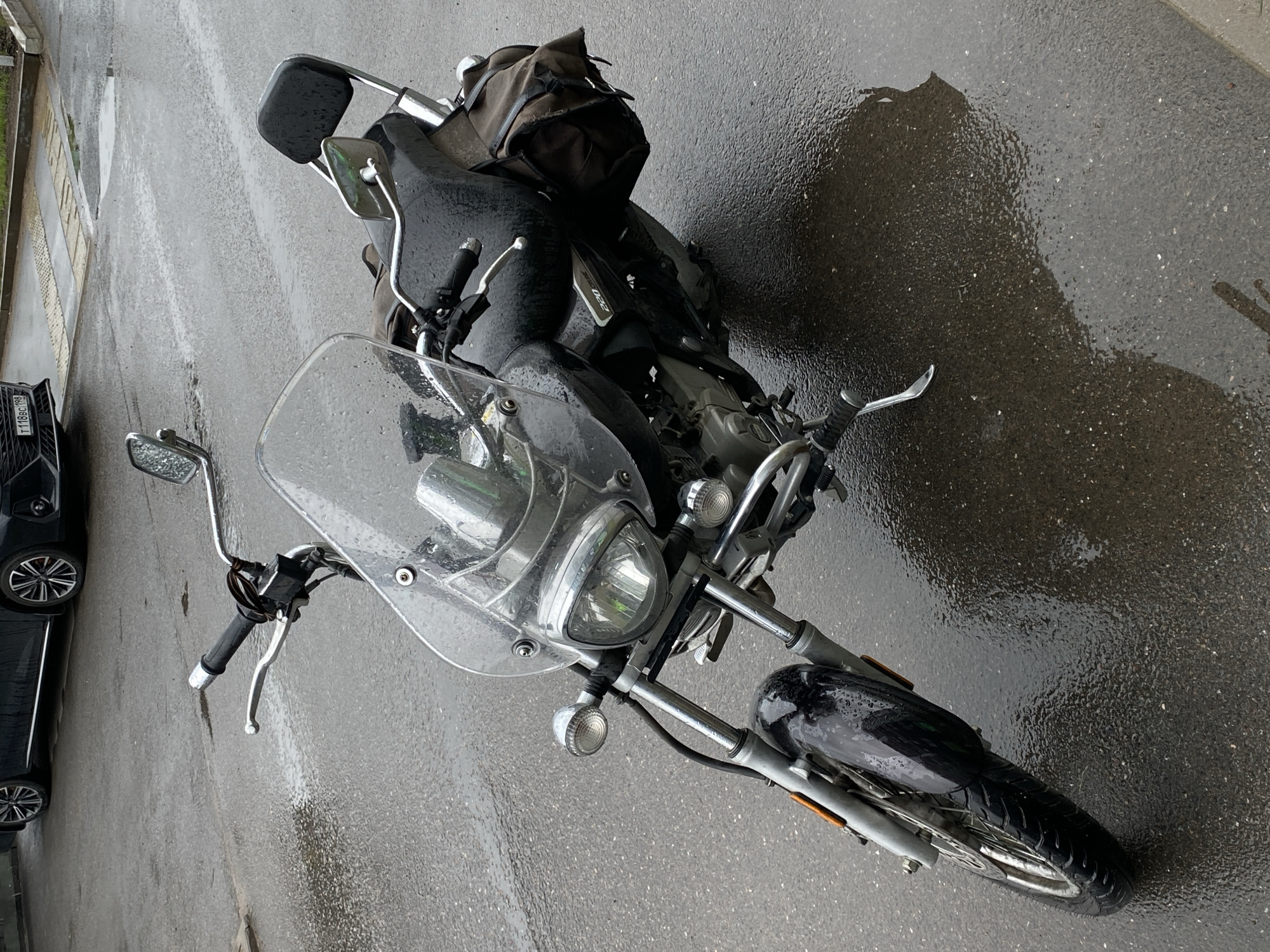 The image shows a motorcycle parked on a wet street in what is likely Saint Petersburg, Russia, based on the vehicle registration plate seen on a black car to the left ("T 118 BC 198"). The motorcycle has a windshield and a bag strapped to the back. The street surface is reflective, indicating recent or ongoing rain.