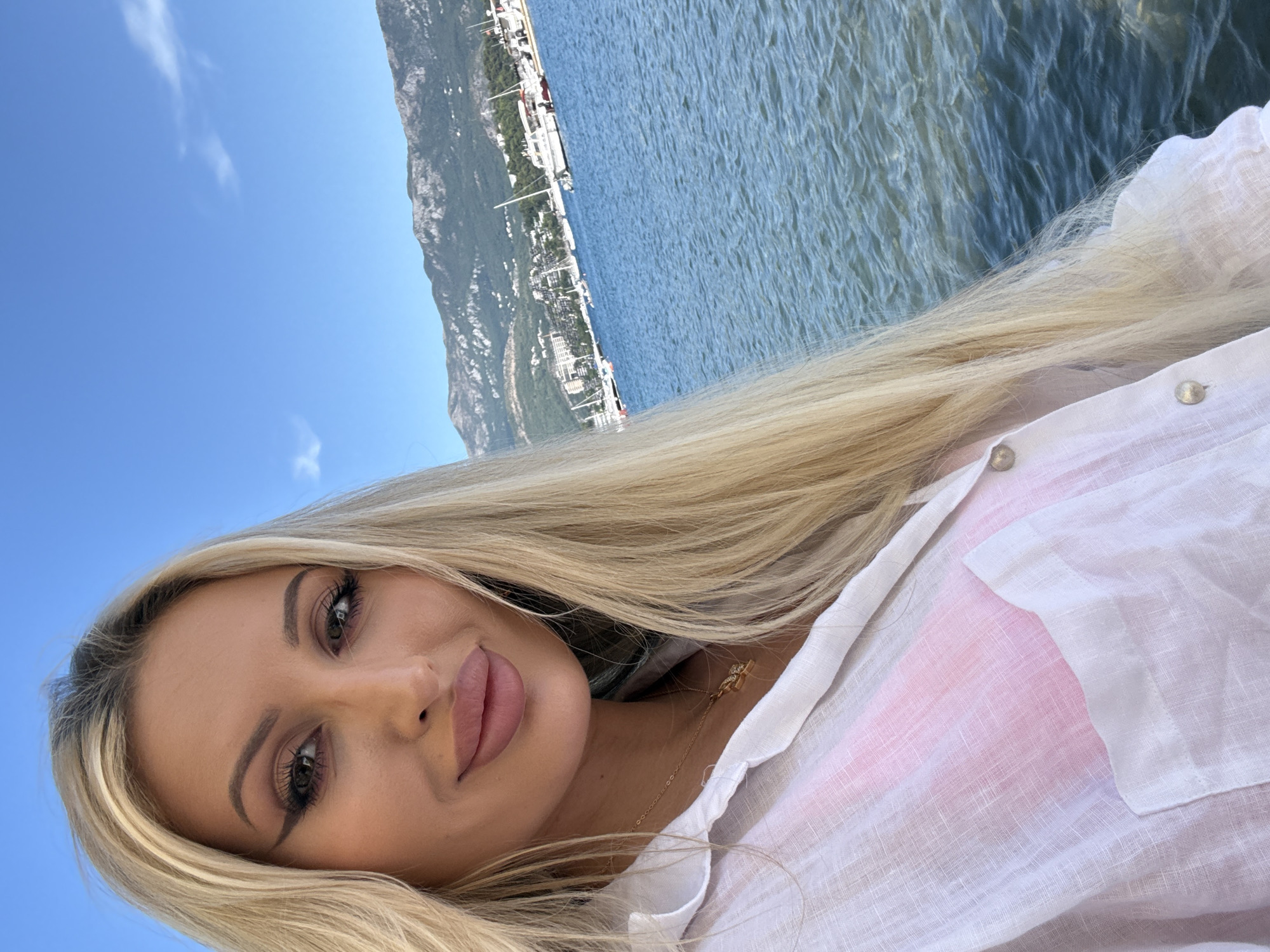 A woman with long blonde hair and full makeup, wearing a white linen shirt over a pink bra, takes a selfie by the sea. She is positioned in the foreground, with the blue water and a mountainous coastline dotted with buildings and boats in the background. The location is identified as Budva, Montenegro. The sky is clear blue with a few wispy clouds.