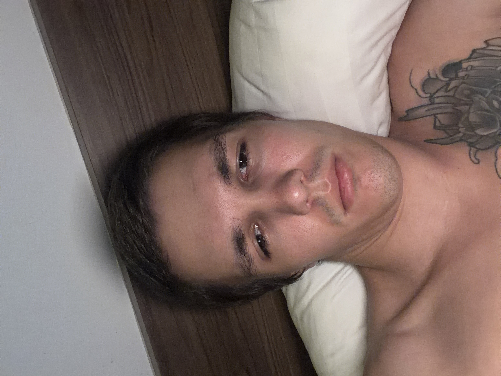 The image shows a close-up, low-angle, selfie-style shot of a young adult male lying on a pillow in bed. He has short brown hair, fair skin, and is shirtless. His eyes are open and looking slightly upwards and to the left of the camera. He has a light mustache and stubble. A tattoo is visible on his chest. The background consists of a pillow and a wooden headboard. The lighting suggests the image was taken indoors, likely in a bedroom. There is no indication of the specific location within the city of Đà Nẵng, Vietnam.