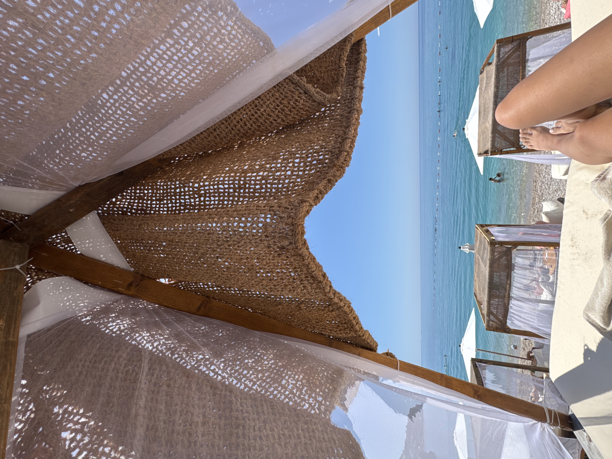 The image captures a first-person perspective from within a rustic beach cabana, providing a view of a serene seaside scene in Vrba, Montenegro. In the immediate foreground, the viewer's tanned bare legs and feet rest on a light-colored cushioned lounger, extending towards the lower right. Overhead, the cabana's roof is constructed from a dark brown, loosely woven natural fiber material, with sections of translucent white mesh fabric interwoven, allowing sunlight to filter through and create dappled light patterns.
Beyond the cabana, the midground reveals a pebbly beach populated with other wooden cabanas featuring white sheer curtains and white beach umbrellas. Several individuals are visible on the beach and wading in the clear, blue-green water near the shore. A distinct line of white buoys or markers stretches horizontally across the ocean, delineating a swimming area. In the background, the calm, expansive sea extends to meet a bright, cloudless blue sky at the horizon. The scene depicts a sunny and tranquil day at the beach.