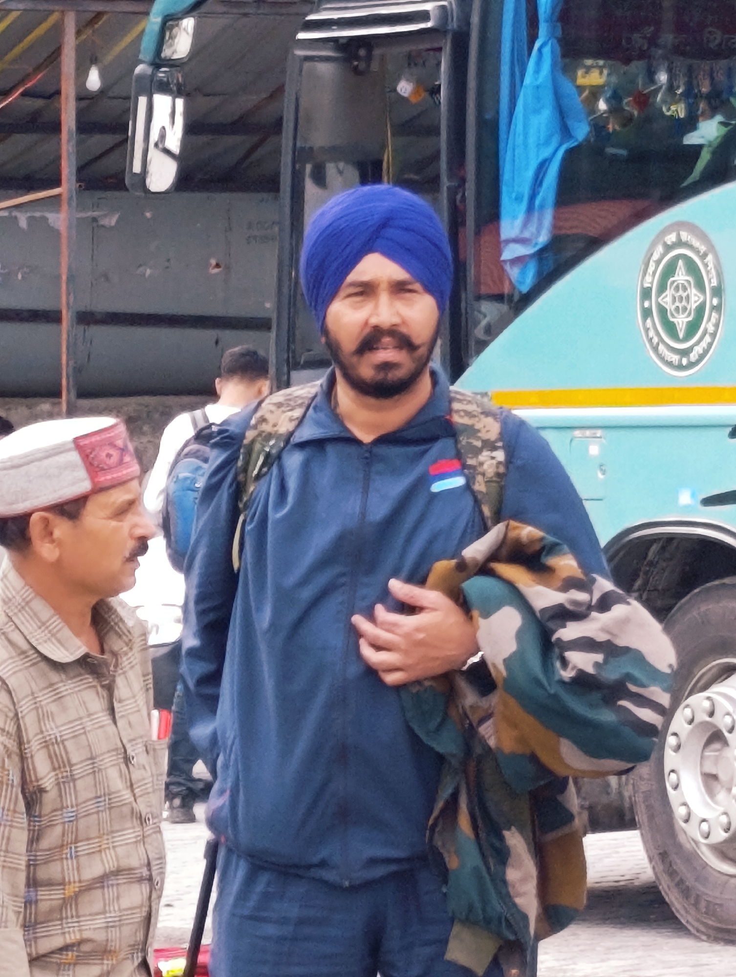 The image shows a group of people near a light blue bus in what appears to be a bus station or parking area in Manali, India. The foreground features two men. The man on the left is wearing a plaid shirt and a traditional Himachali cap. The man on the right is wearing a blue turban, a blue jacket, and a camouflage pattern over his arm. Behind them, other individuals and a blue bus are visible. The environment suggests an outdoor setting, possibly in a mountainous region given the mention of Manali.
