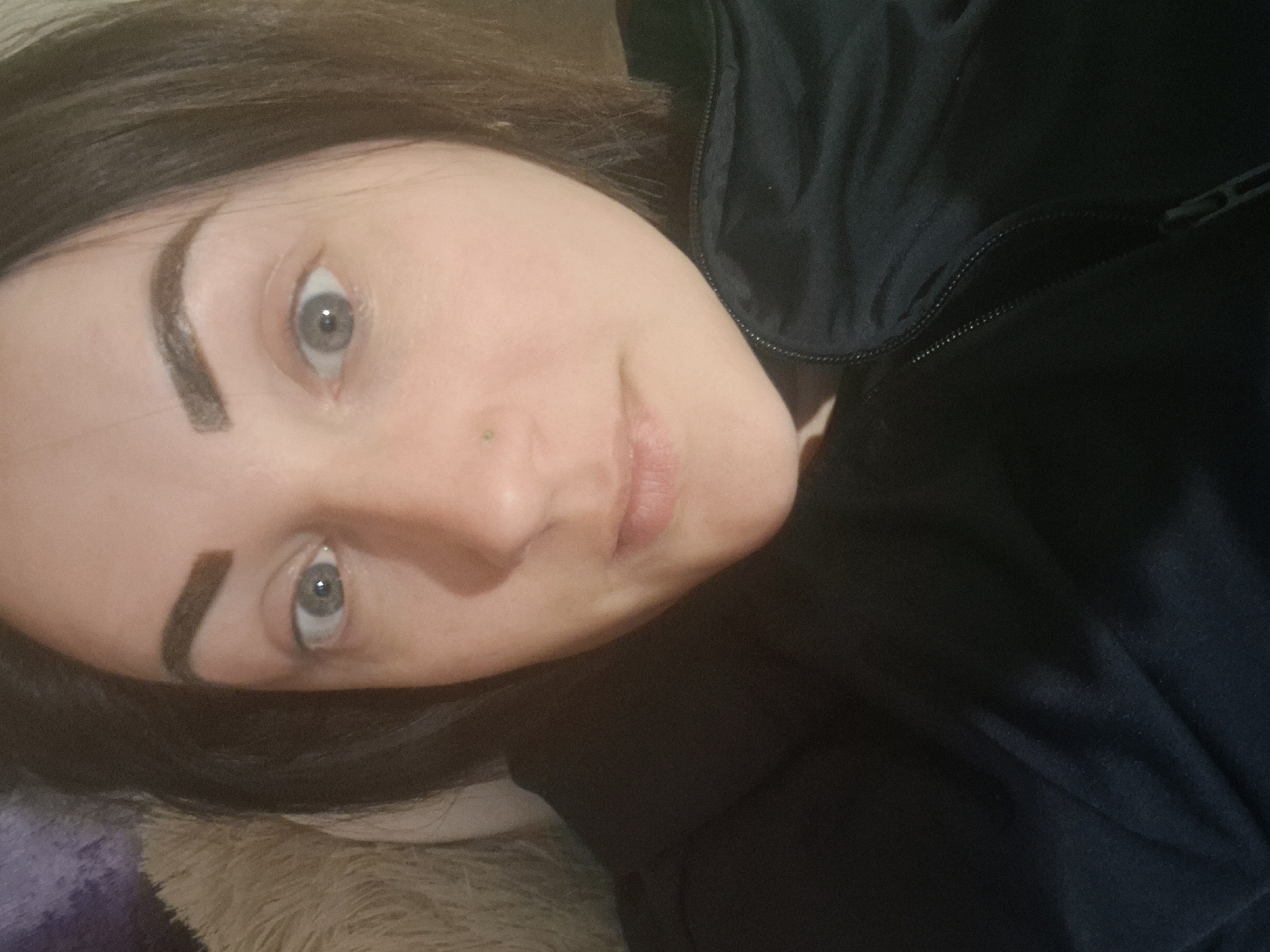 The image is a close-up selfie of a woman's face, taken from a slightly high angle. She has dark hair, fair skin, and light blue eyes. Her eyebrows are dark and well-defined, and she appears to be wearing light makeup. There is a small, greenish nose stud visible on her left nostril. She is wearing a dark-colored zippered jacket. The background is blurred, with hints of a furry, textured surface on the left side and a dark, wrinkled fabric on the right. The lighting suggests an indoor setting. This image does not provide any information that would allow for the determination of its location in the city of Sandwell, United Kingdom.