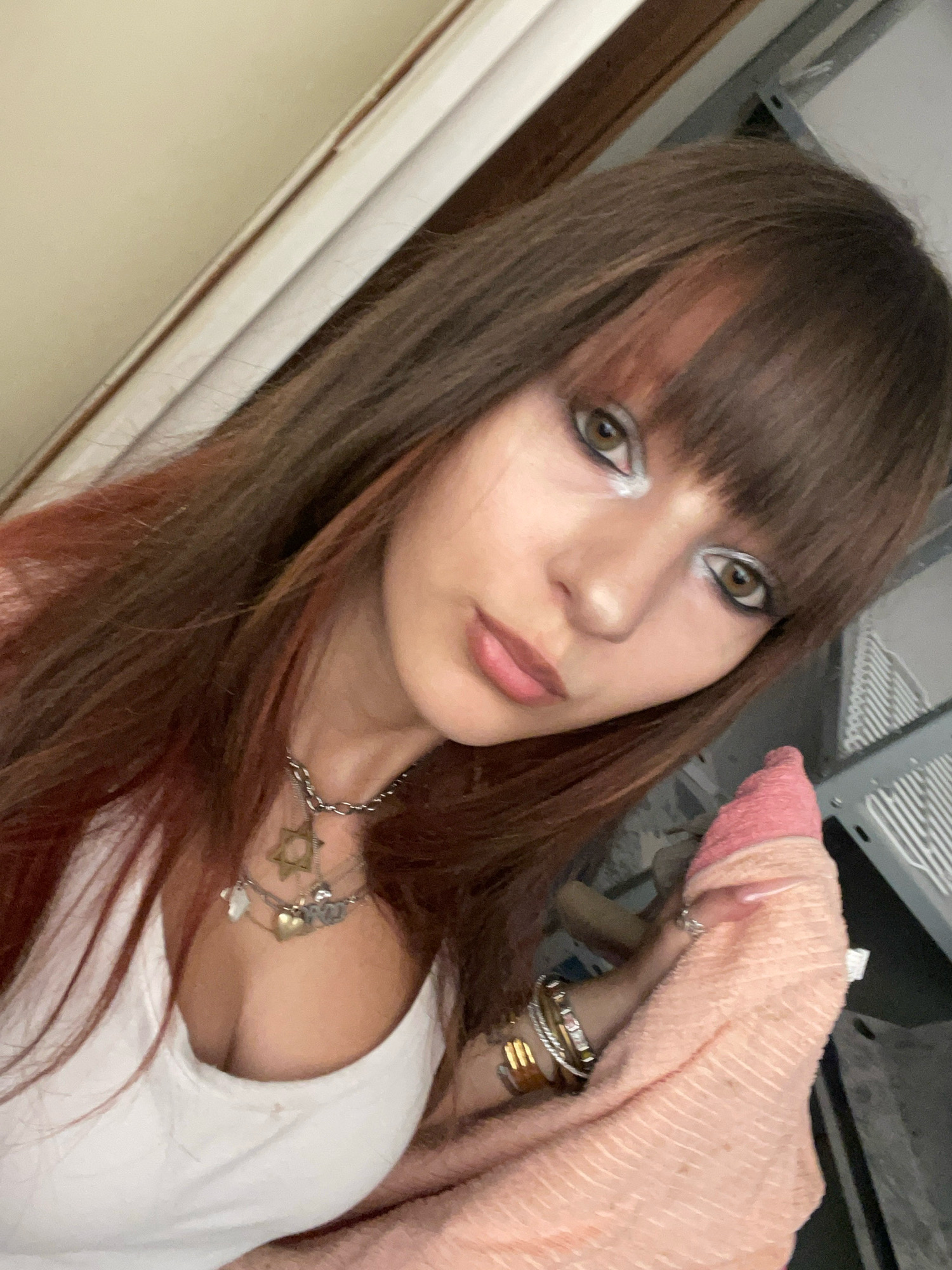 A young woman with long, brown hair with reddish highlights and bangs is shown in a selfie. She is wearing a white tank top and several necklaces, one of which appears to be a Star of David. Her makeup includes heavy eyeliner and shimmery eyeshadow. She is holding a pink towel with her right hand, and on her left hand, she is wearing several bracelets, including a gold one. In the background, a metal structure with white grates is visible, suggesting an indoor setting. The overall lighting is dim, with a warm tone. The location is the city of Raanana, Israel.