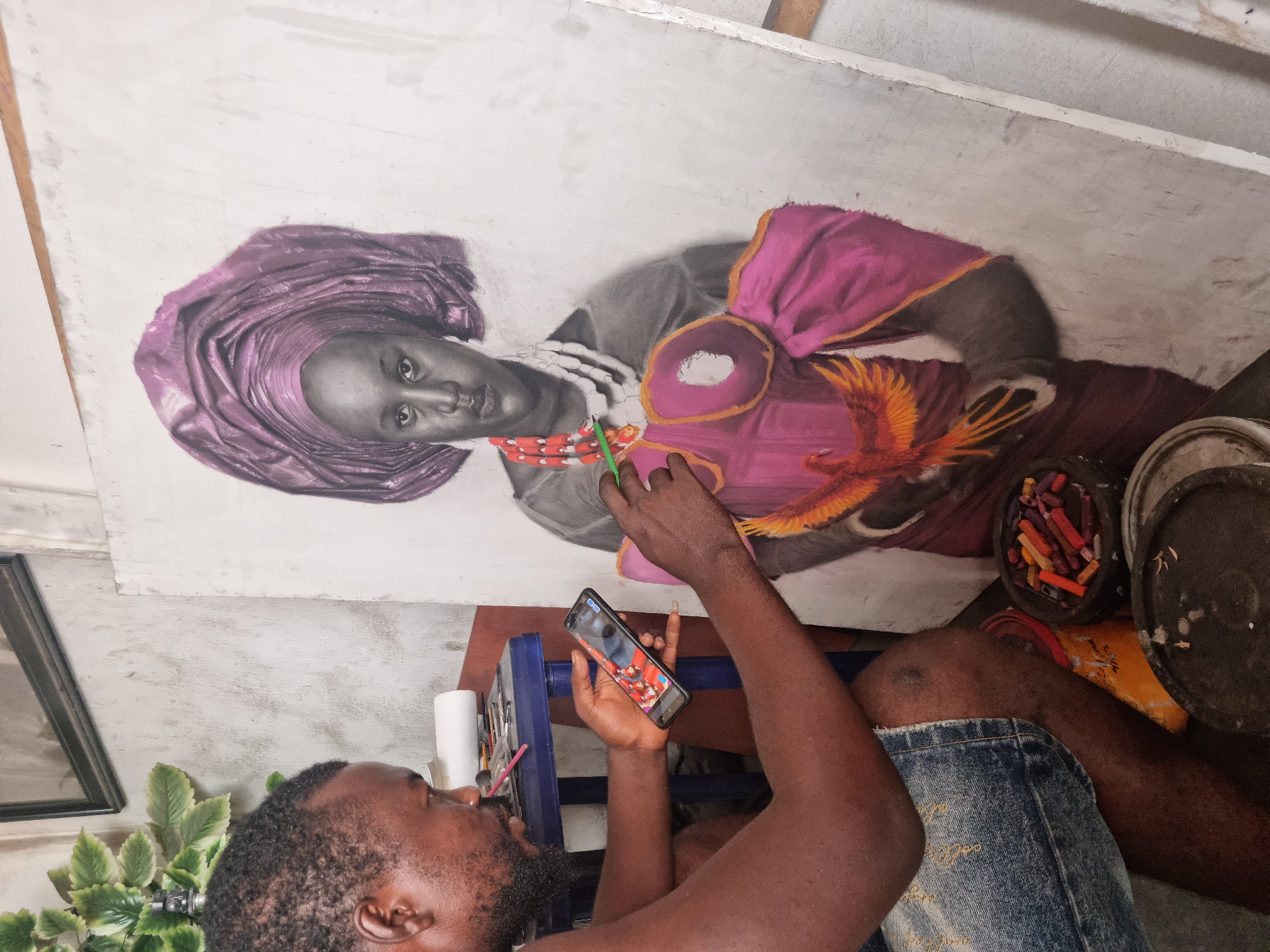 In an art studio in Onitsha, Nigeria, a Black male artist is depicted from an overhead angle, immersed in his work. He is seated or kneeling, wearing denim shorts, with his gaze directed towards a smartphone held in his left hand. His right hand holds a green-handled tool, possibly a brush or blending stump, and is actively working on a large portrait laid out on a flat surface in front of him.
The artwork features a lifelike portrait of a Black woman, rendered with a combination of grayscale tones for her skin and vibrant colors for her attire. She wears a striking purple headwrap (gele) and a pink outfit adorned with orange accents and a decorative element resembling a colorful bird or feathers on her chest. The portrait appears to be in progress, with some background areas still unpainted.
Beside the artist, various art supplies are visible, including a container filled with colorful crayons or pastels, and other brushes. Artificial green plant leaves are also seen near the artist's left side, alongside a partially visible framed picture. The artist has a beard and short dark hair, and his expression suggests focus as he references his phone, which displays a colorful image, likely a part of his artistic process or a reference photo. The setting is an indoor workspace, indicated by a light-colored floor and simple background. No discernible text is visible within the image beyond what might be on the smartphone screen.