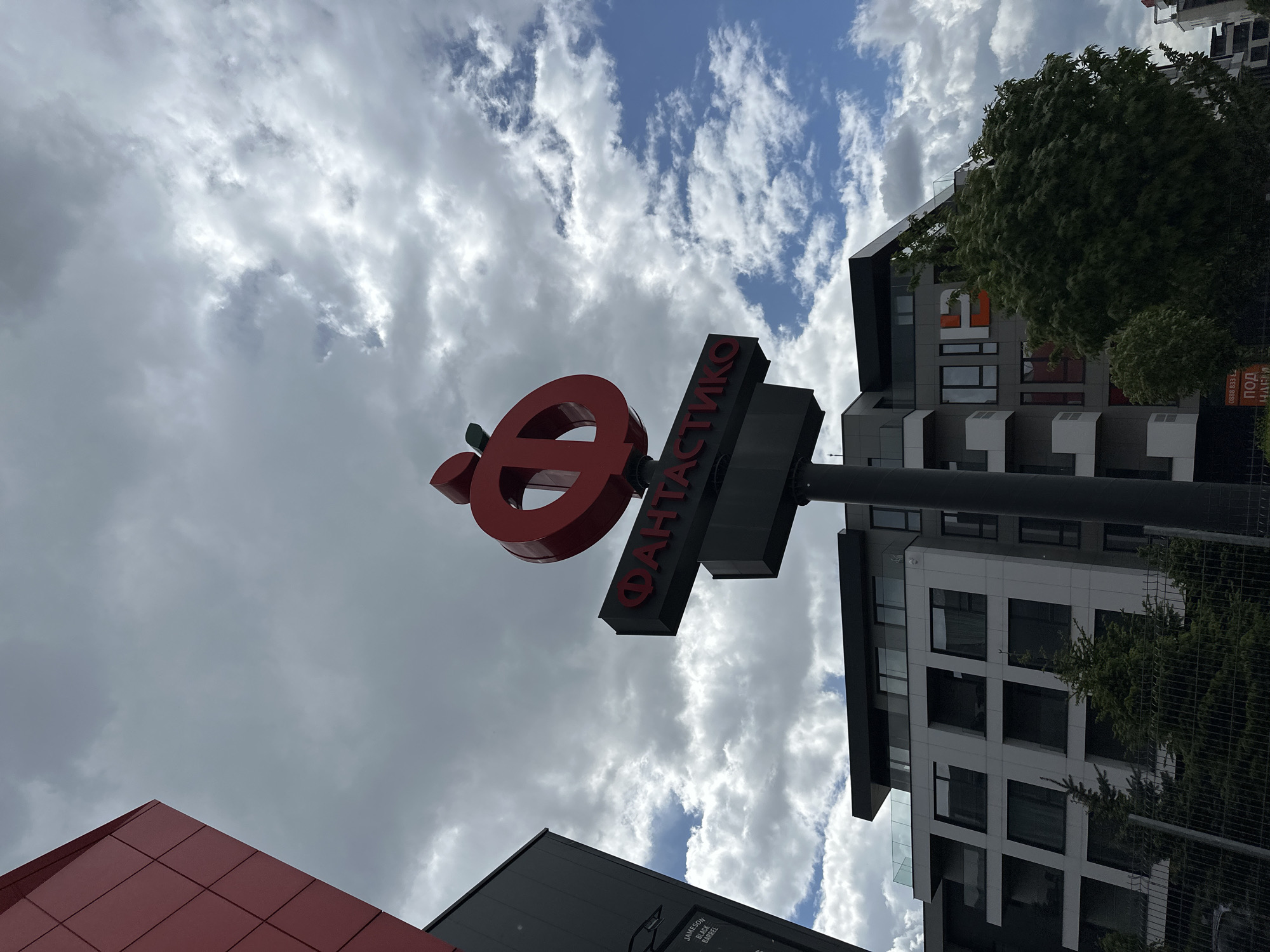 The image shows the exterior of a Fantastico supermarket in Sofia, Bulgaria. The red Fantastico logo, a stylized apple forming the letter "O," is prominently displayed against a backdrop of cloudy sky. The Cyrillic text "ФАНТАСТИКО" (Fantastico) is visible on the sign below. Modern apartment buildings with mixed gray, black, and white facades stand in the background, partially obscured by trees. A red-paneled wall is visible in the lower left corner of the frame. The scene is shot from a low angle, looking upwards at the sign and buildings.