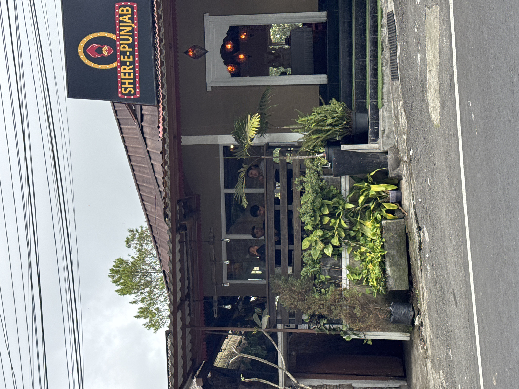 In Kintamani, Indonesia, a two-story establishment features a restaurant named "SHER-E-PUNJAB" with a prominent sign. The building has a rust-colored corrugated metal roof and a dark brown facade. A covered porch area on the second level overlooks the street and is furnished with a wooden railing. Several people are visible behind the railing, appearing to be patrons. Decorative pendant lights hang from the ceiling of the porch. The entrance to the building is at street level, marked by a white archway with multiple orange lanterns illuminating the interior, where a statue is visible. Greenery, including potted plants and vines, surrounds the building at the base, and a paved pathway with steps leads to the entrance. A street with a white line marking is in the foreground, and overhead power lines are visible against a cloudy sky.