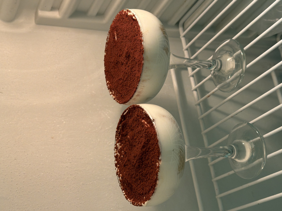 Two glasses of tiramisu rest sideways on a white refrigerator shelf. Each glass contains a white creamy base topped with a generous layer of cocoa powder. The glasses have long stems and round bases. The wire racks of the refrigerator shelf are visible behind the glasses, and the smooth white interior walls of the fridge are adjacent to them.