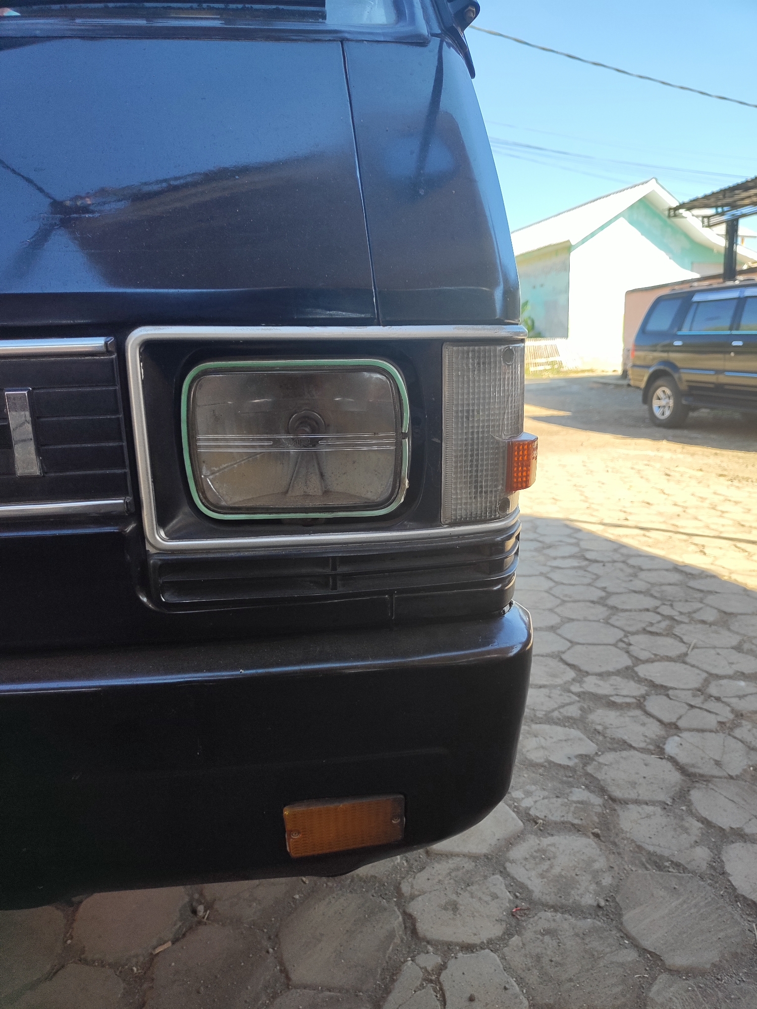 In the city of Pujerbaru, Indonesia, a close-up, low-angle shot captures the front right corner of a dark-colored vehicle, possibly a van. The vehicle's headlight, framed by a silver and green bezel, is visible and appears to have some damage. To the right of the headlight, a clear turn signal with an amber bulb is mounted. Below the headlight and turn signal, a horizontal grille is present. A small, rectangular amber reflector is located on the lower front bumper of the vehicle. The vehicle is parked on a paved surface with a hexagonal pattern. In the background, a light-colored building and a black SUV are visible under a clear blue sky. The sun casts shadows on the ground.