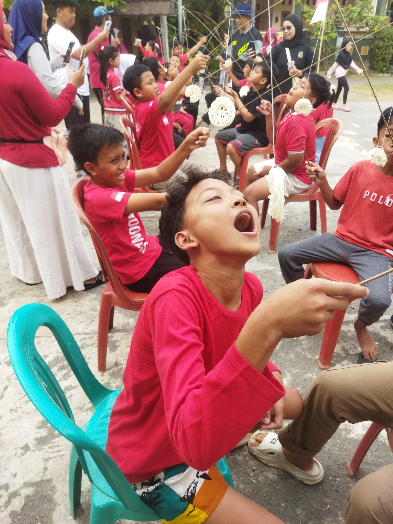 The image captures an outdoor event in Banjar, Indonesia, likely a community gathering or festival. The foreground features several children engaged in a game where they attempt to eat crackers suspended from strings. One boy, wearing a red shirt and colorful shorts, is prominently positioned, mouth wide open, reaching for a cracker with a stick. To his right, another boy in a red shirt and grey pants is also participating in the game. Behind them, more children and adults are visible, some holding phones, suggesting they are either spectators or documenting the event.
The environment appears to be a paved outdoor area, possibly a street or a courtyard, with some vegetation and buildings in the background. The participants are dressed in casual attire, with many wearing red shirts, which could indicate a team or a theme for the event. The overall atmosphere seems lively and festive, with a focus on a traditional game. The presence of Indonesian flags further confirms the location and cultural context of the scene.