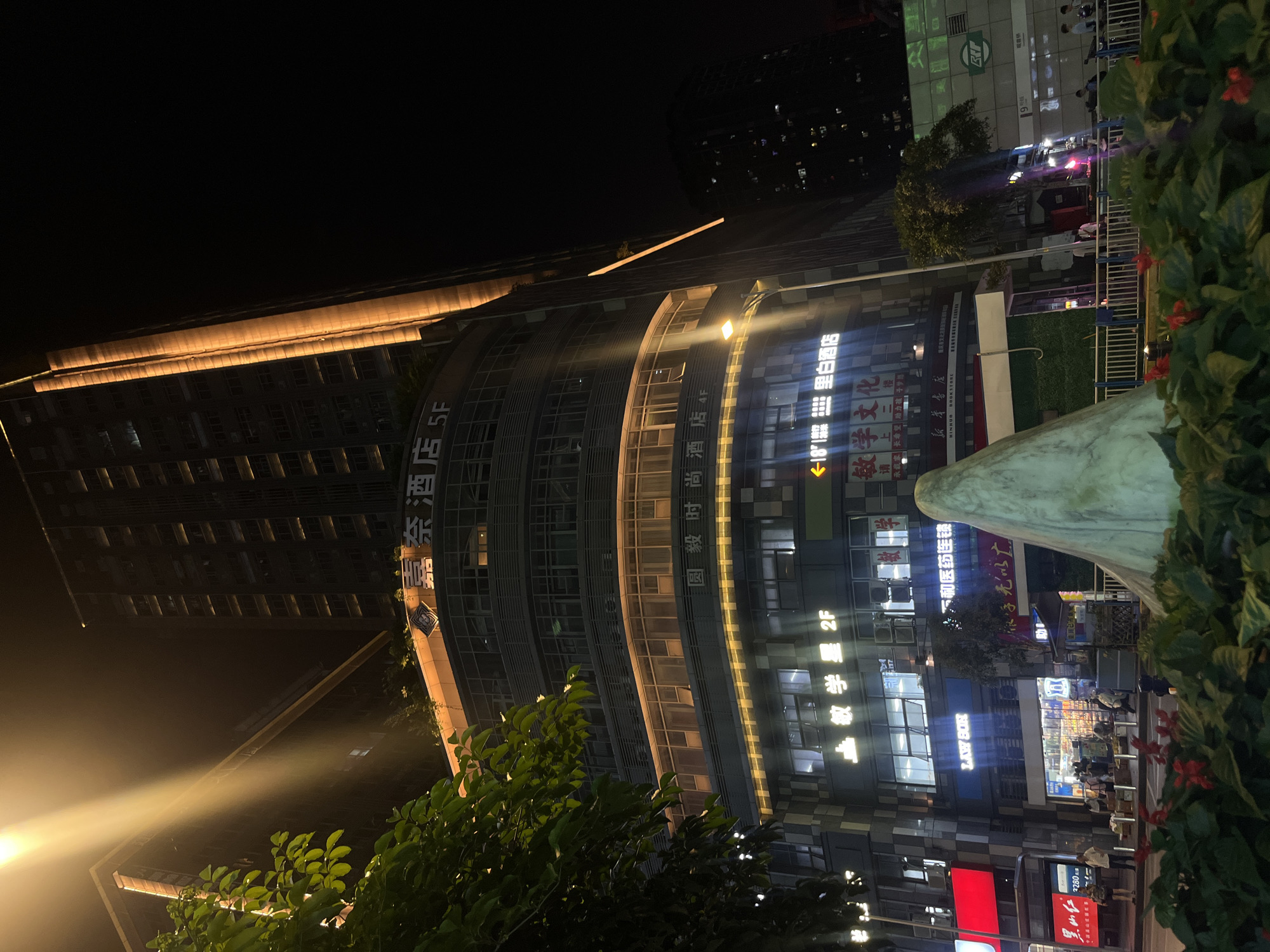 This nighttime image captures a lively urban scene in Liangjiang, China, characterized by brightly lit buildings and commercial activity. The setting appears to be a busy street or plaza lined with multi-story establishments.
In the foreground, lush green foliage, including bushes with small red flowers, partially obscures the view. To the right, a large, light-colored, abstract sculpture or monument stands out. Beyond the greenery, several modern buildings dominate the frame. One prominent building features a distinctive curved glass facade, illuminated by strips of golden light. Another taller building in the background has rows of rectangular windows.
The scene is bustling with activity typical of an urban night. People can be faintly seen moving about at street level, some near storefronts, while others are on a walkway. Artificial light sources, including streetlights and illuminated building signs, cast strong beams and reflections, highlighting the architectural details and creating a vibrant atmosphere.
Visible text on the building signs provides specific location cues: "壹奈酒店 5F" (Yi Nai Hotel 5F), "圆毅时尚酒店 4F" (Yuanyi Fashion Hotel 4F), "里白酒店" (Libai Hotel), "敏学文化" (Minxue Culture), and "新華書店" (Xinhua Bookstore), which also has "HINHUA BOOKSTORE" in English. A "LAWSON" convenience store sign is also prominent. Other texts include "敏学星 2F", "上二楼" (Upstairs 2nd Floor), "承载家长希望助力孩子梦想" (Carrying parents' hopes, helping children's dreams), and floor indicators like "5F", "4F", "2F". The presence of these businesses suggests a commercial and cultural hub. The time of day is clearly night, with no indications of adverse weather.